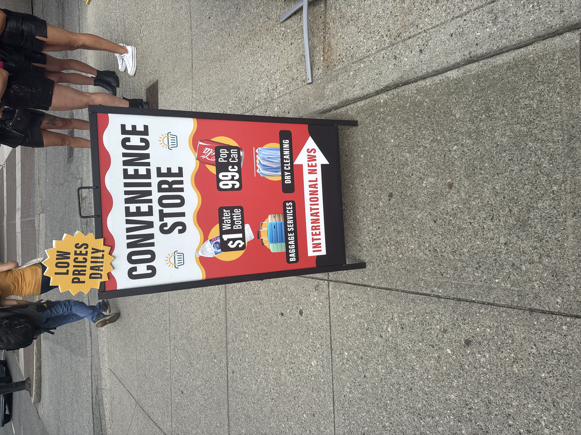 This is an eye-level, medium shot of a sandwich board sign for a convenience store. The sign is located on a concrete sidewalk, with a street visible in the background. Several people are walking in the background, and their legs are visible. The sign has a red and white color scheme and lists various items and services offered by the store, including "LOW PRICES DAILY," "CONVENIENCE STORE," "$1 Water Bottle," "99c Pop Can," "BAGGAGE SERVICES," "DRY CLEANING," and "INTERNATIONAL NEWS." The sign is propped up by metal legs. A grey metal pole extends from the right side of the frame. The environment appears to be an urban setting.