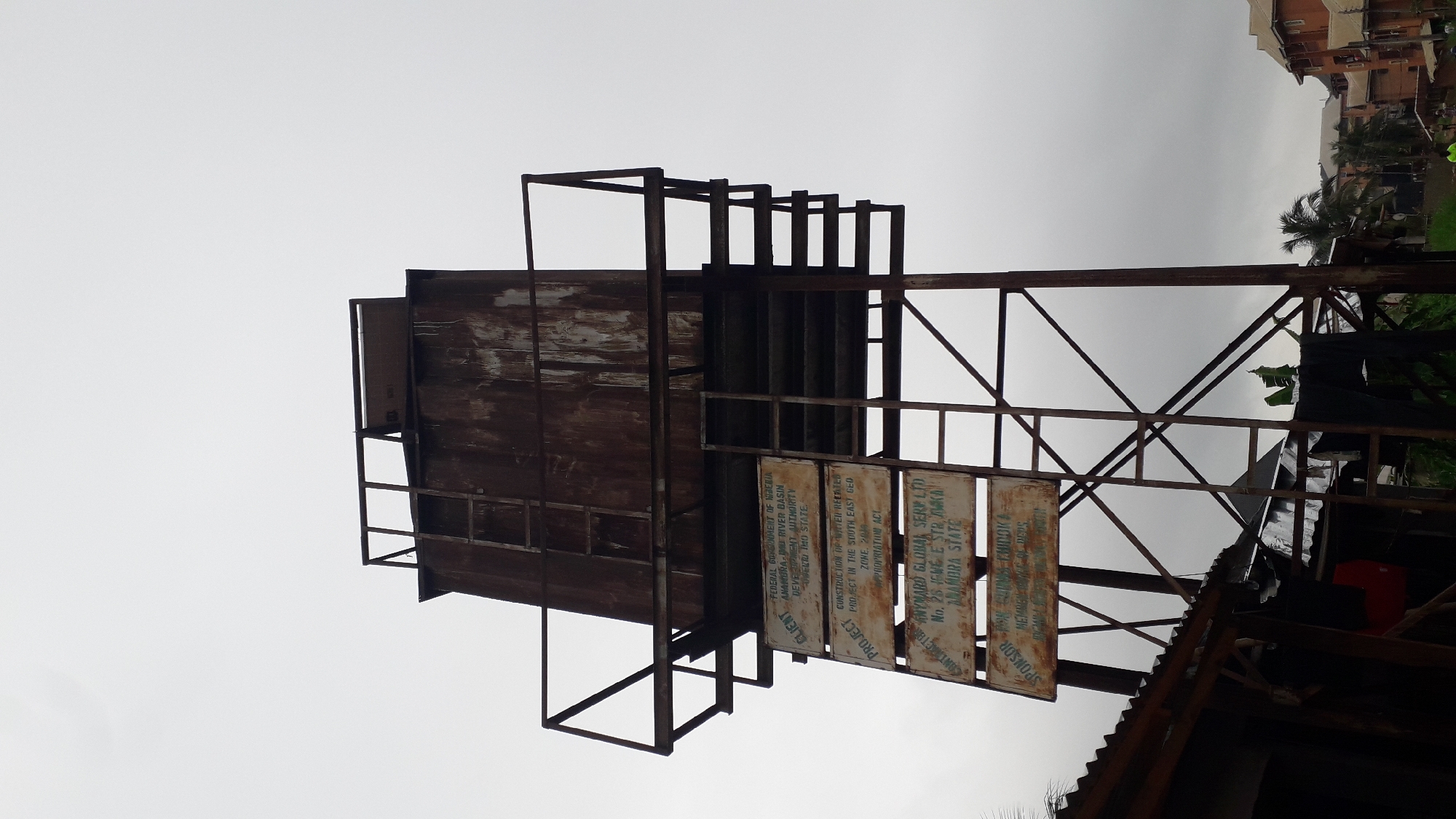 The image shows a rusted, elevated water tank in Nkpor, Nigeria. The tank is supported by a metal frame and has weathered wooden sides. Three signs are attached to the structure. The text on the signs indicates the tank was part of a project by the Federal Government of Nigeria, the Anambra-Imo River Basin Development Authority, and Hayward Global Serv LTD. Buildings are visible in the background. The weather appears overcast.