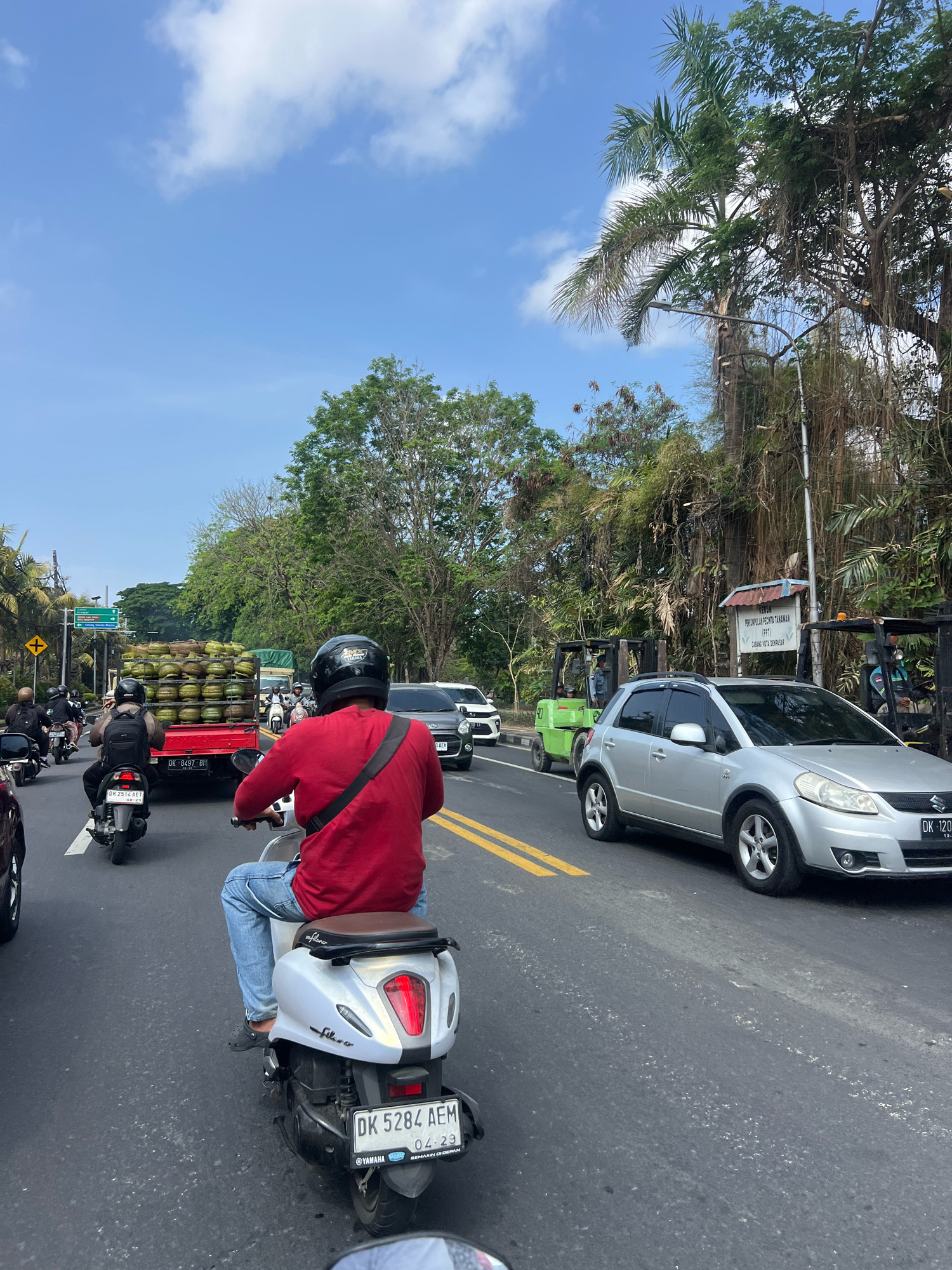 This is a street scene in Denpasar, Indonesia. The image captures a traffic jam on a sunny day with a blue sky and some clouds. In the foreground, a person is riding a silver scooter, wearing a red long-sleeved shirt and blue jeans. Another scooter, black in color, is behind them. Further up the road, a red truck is carrying a large load of coconuts. Several other vehicles, including a silver Suzuki car and two green forklifts, are also present on the road. Lush green trees and vegetation line the sides of the street. A signpost is visible in the background, partially obscured by trees and hanging vines. The presence of multiple scooters and the style of the vehicles and signage suggest a location within a Southeast Asian urban environment.