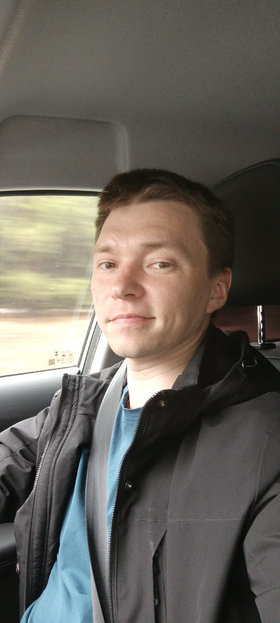 A medium-shot captures a Caucasian male inside a car. He is wearing a black jacket over a blue T-shirt and a seatbelt. His hair is short and brown, and he looks directly at the camera with a slight smile. Outside the car window, blurred foliage suggests movement, indicative of the car being in transit. The interior of the car includes grey upholstery and a black headrest. The city of Niva, Russia is not discernible from the image.