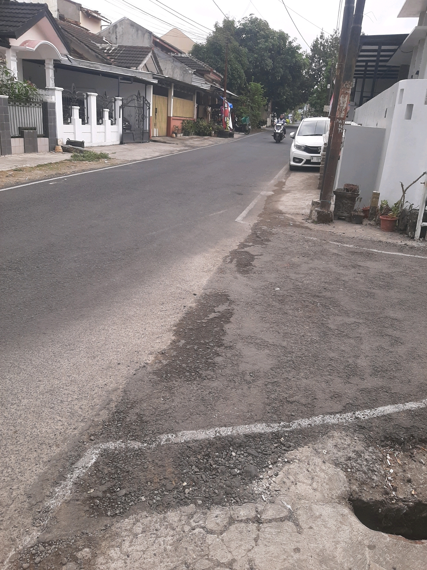 The image depicts a street scene in Sumampir, Indonesia, on an overcast day. The foreground shows a section of asphalt road with a distinct line of white paint marking the edge. Adjacent to the road is a damaged concrete drainage channel, with loose gravel and debris visible. Further into the scene, a white car is parked on the side of the road. Behind the car, a motorcycle with two occupants is moving away from the camera. The street is lined with houses, some with tiled roofs and white fences, and interspersed with lush green trees. A utility pole stands prominently on the right side of the road. The overall impression is of a typical residential street in a tropical urban environment.