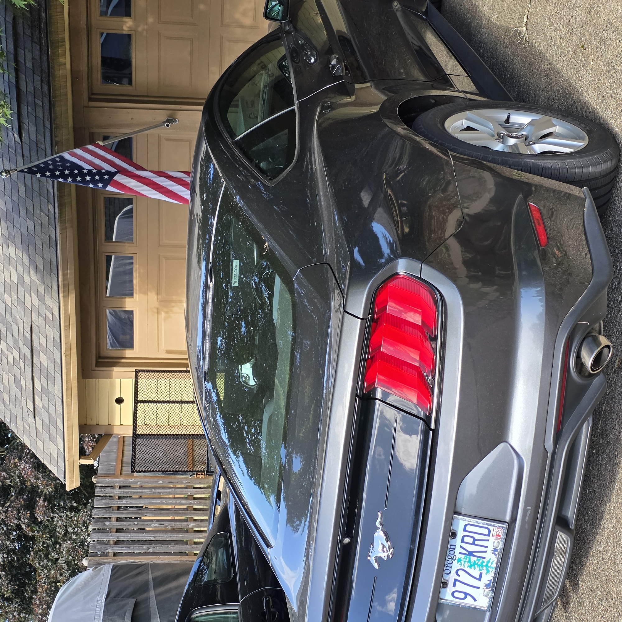 The image depicts the rear passenger side of a dark gray Ford Mustang parked on an asphalt driveway in front of a detached garage. The garage has beige double doors with windows and a pitched roof with gray shingles. An American flag is mounted on the right side of the garage. A wooden privacy fence is visible to the left of the garage. Next to the Mustang, another black car is partially visible. The Mustang has an Oregon license plate that reads "972 KRD". The scene is likely in Beaverton, United States of America, given the Oregon license plate.