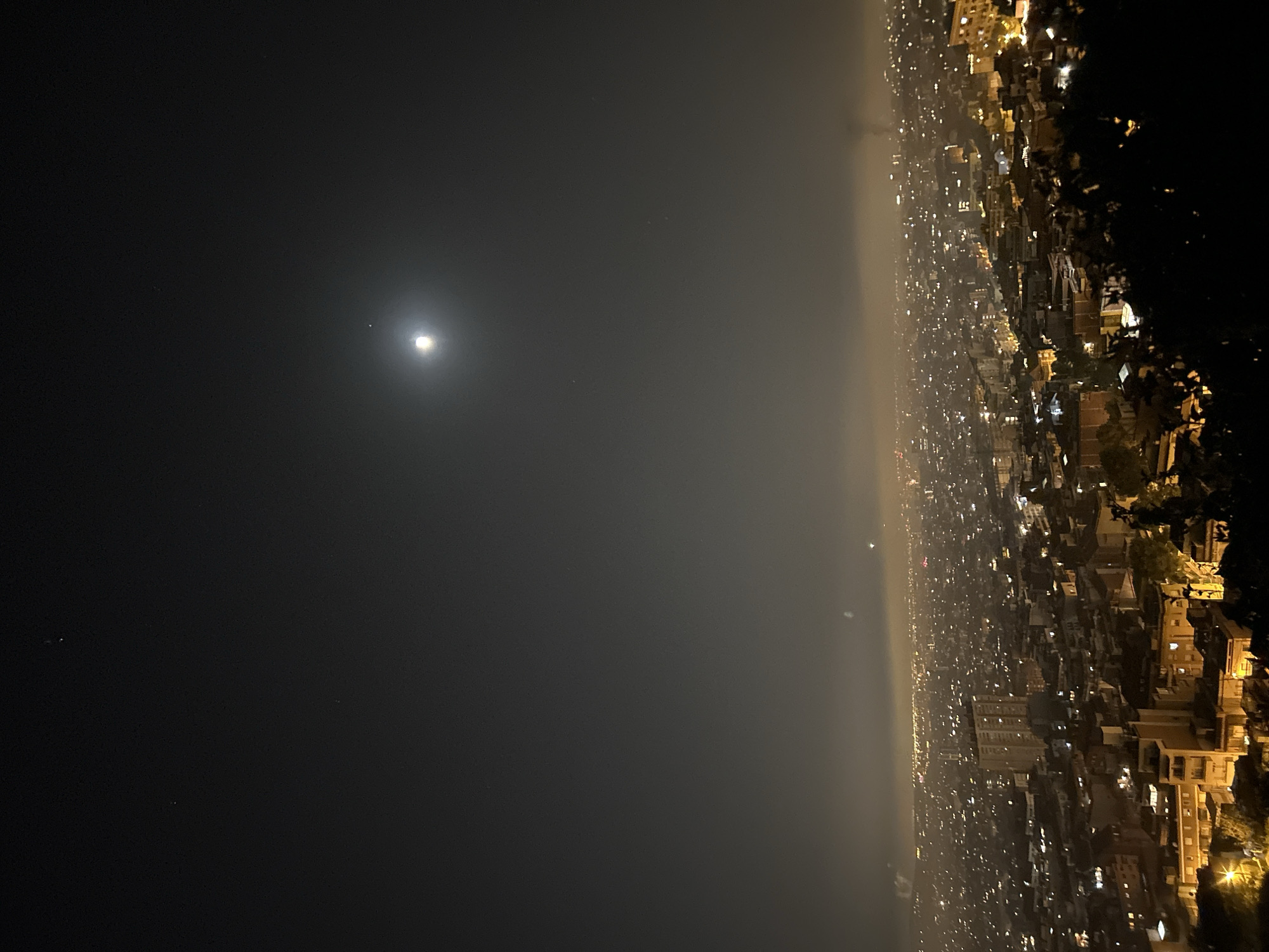 An elevated nighttime panoramic view of Barcelona, Spain, showcases a sprawling cityscape illuminated by countless lights. The urban expanse stretches into the distance, veiled by a hazy atmosphere that softens the glow of the city. Above, a bright moon shines prominently in a dark sky, accompanied by a few distant stars. The foreground is dominated by dense foliage, silhouetted against the illuminated city, hinting at a viewpoint from a hill or elevated park overlooking the metropolis.