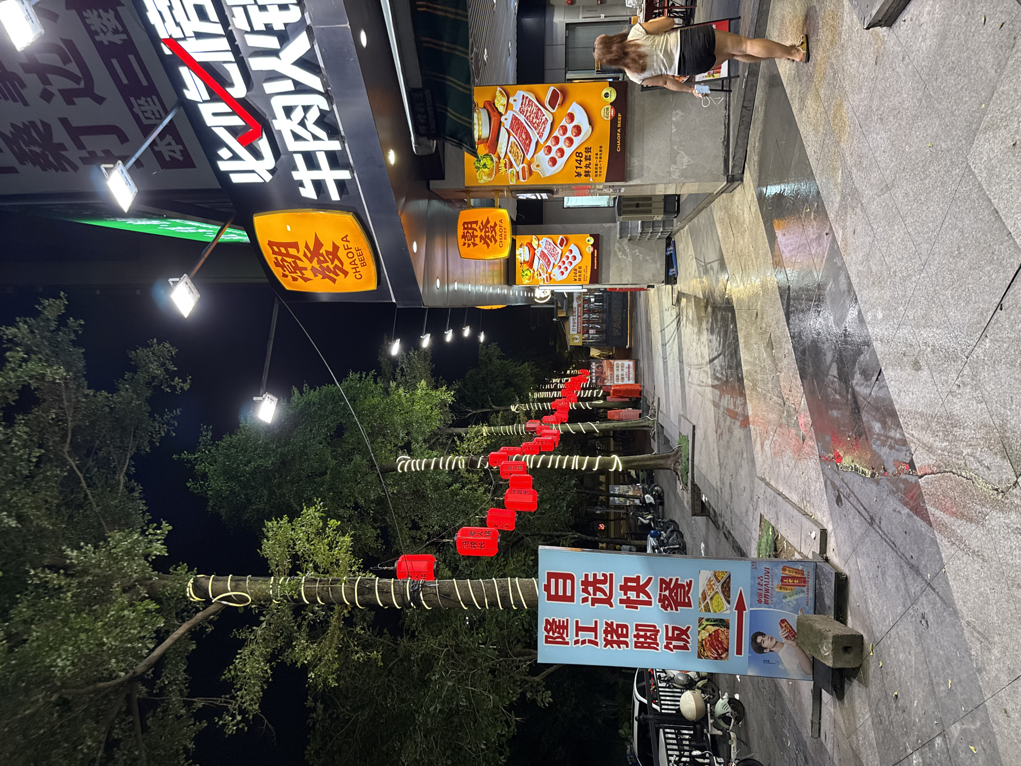 This media file captures a nighttime scene on a street in Haizhu District, China. The focus is on a bustling street-level commercial area with multiple food establishments. The foreground features trees adorned with white string lights and red lanterns, creating a festive atmosphere. A prominent sign on the right advertises "隆江猪脚饭" (Longjiang Pig Trotter Rice) and "自选快餐" (Self-service Fast Food), along with images of food and a smiling young man.
Further down the street, illuminated signs for "CHAOFA BEEF" are visible, showcasing images of beef dishes and prices, such as a "鲜丸套餐" (Fresh Ball Set Meal) for ¥148. The street itself appears to be paved with stone, and reflections on the wet surface suggest it might have rained recently or the street is being cleaned.
A few individuals are present, including a woman with long, light-colored hair wearing a white t-shirt and dark shorts, walking away from the camera. Another person is partially visible further down the street. Motorcycles are parked along the side of the road, indicating this is a common mode of transportation. The overall impression is one of an active, commercial street at night, with a mix of traditional and modern elements, likely a popular spot for dining and shopping. The lighting and decorations create a lively and inviting ambiance.