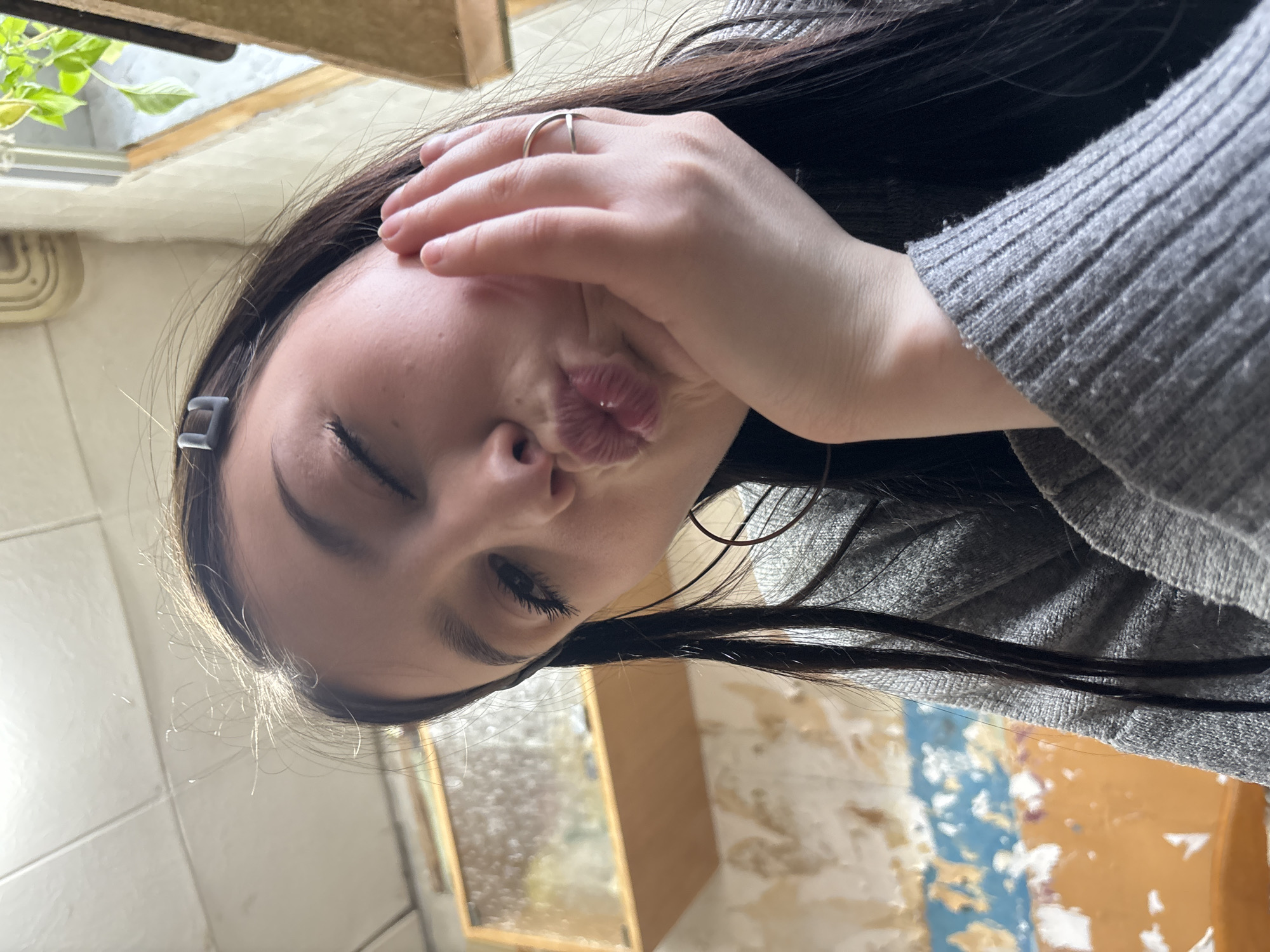 The image, rotated 90 degrees counter-clockwise, presents a close-up portrait of a young woman with long, dark hair. She is captured in a playful pose, with her left eye closed or squinting, and her lips pursed in a "kiss" or "duck face" expression. Her right hand is brought up to her cheek, partially covering her face, and a silver ring is visible on her ring finger. She is wearing a grey, ribbed long-sleeved sweater. A small, grey rectangular hair clip is clipped into her hair on the left side of her head.
The background is a mix of indoor and outdoor elements. In the upper left (when viewed upright), green leaves of a plant are visible through what appears to be a window or skylight, suggesting natural light and an outdoor view. The lower right background shows an interior wall with peeling paint or wallpaper, featuring patches of light beige, blue, and white, implying a somewhat distressed or informal setting. A blurry rectangular object, possibly a mirror or picture frame, is also discernible. The lighting appears natural, accentuating highlights in her dark hair. The overall impression is one of a lighthearted and candid moment. There is no visible text. The location context for this media is Фоки, Russia.