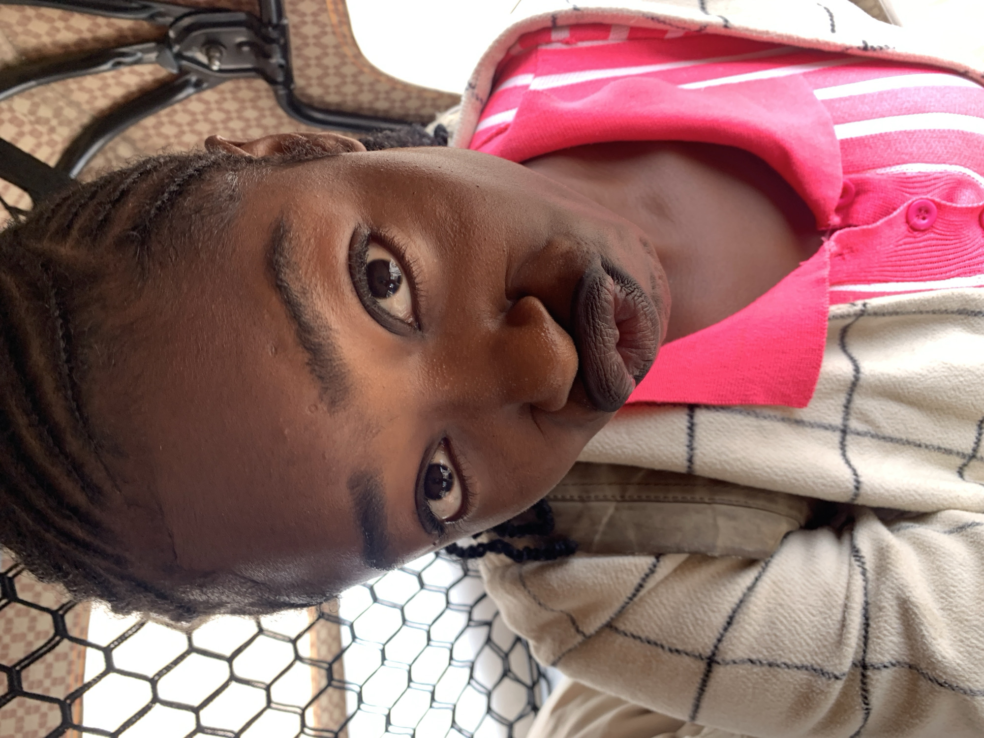 A close-up portrait captures a young female subject with a dark complexion, situated in Rafin Par, Nigeria. Her dark hair is styled in tightly braided cornrows. She has dark eyes, accentuated with eyeliner, fixed directly on the viewer, and her full lips are deliberately pursed into a pout, coated with a dark, matte lip color. She is dressed in a vibrant pink collared shirt with prominent vertical white stripes and a single visible pink button, worn beneath an outer garment of a light beige or off-white color featuring a dark, gridded, or plaid pattern. The background on the left displays a black hexagonal mesh screen with a bright light source beyond it. The background on the right reveals a brown fabric with a repeating lighter geometric pattern, consistent with interior upholstery, alongside dark, angular metallic structural elements. The scene is illuminated by natural light.