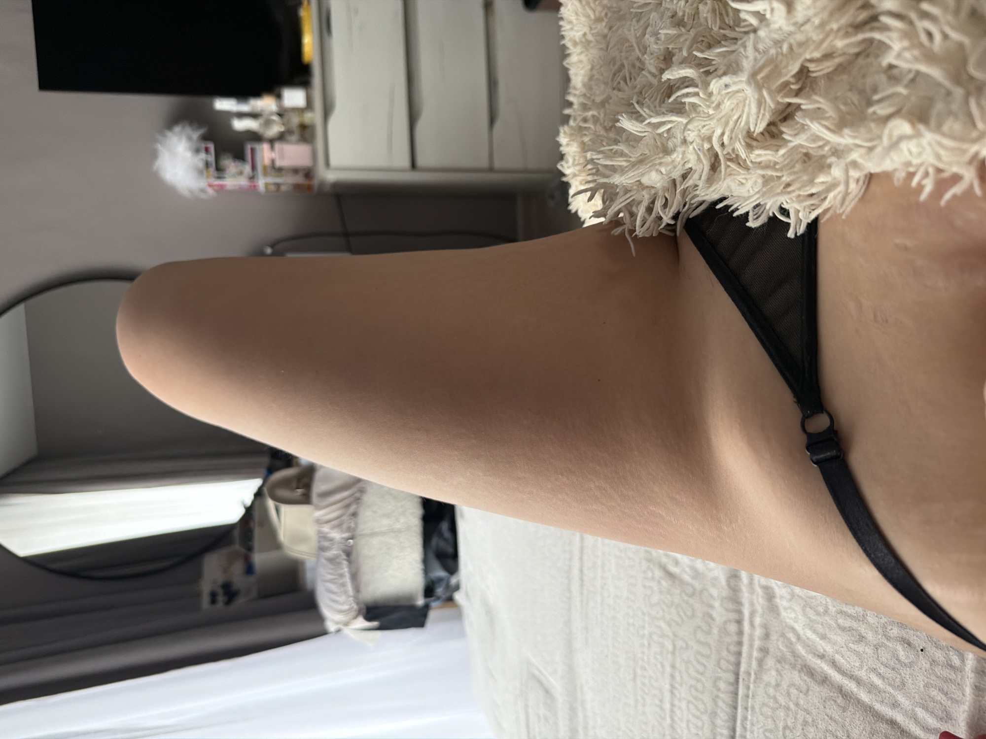 The image displays the bare leg and hip of an individual, reclined on a bed within an indoor room. A black thong-style undergarment with a side adjuster is visible on the hip. A textured, off-white, shaggy blanket partially covers the lower abdomen. The background features a light grey wall. To the left, a window is dressed with white sheer and grey opaque curtains, and a large, dark-framed circular mirror reflects part of the room and window. To the right, a white multi-drawer dresser is visible, topped with various items including bottles, a white feathery decoration, and a partial view of a dark television screen. The bed is covered by a light-colored, textured duvet. The specific location within Chaikovsky, Russia, is not identifiable from the provided visual evidence.