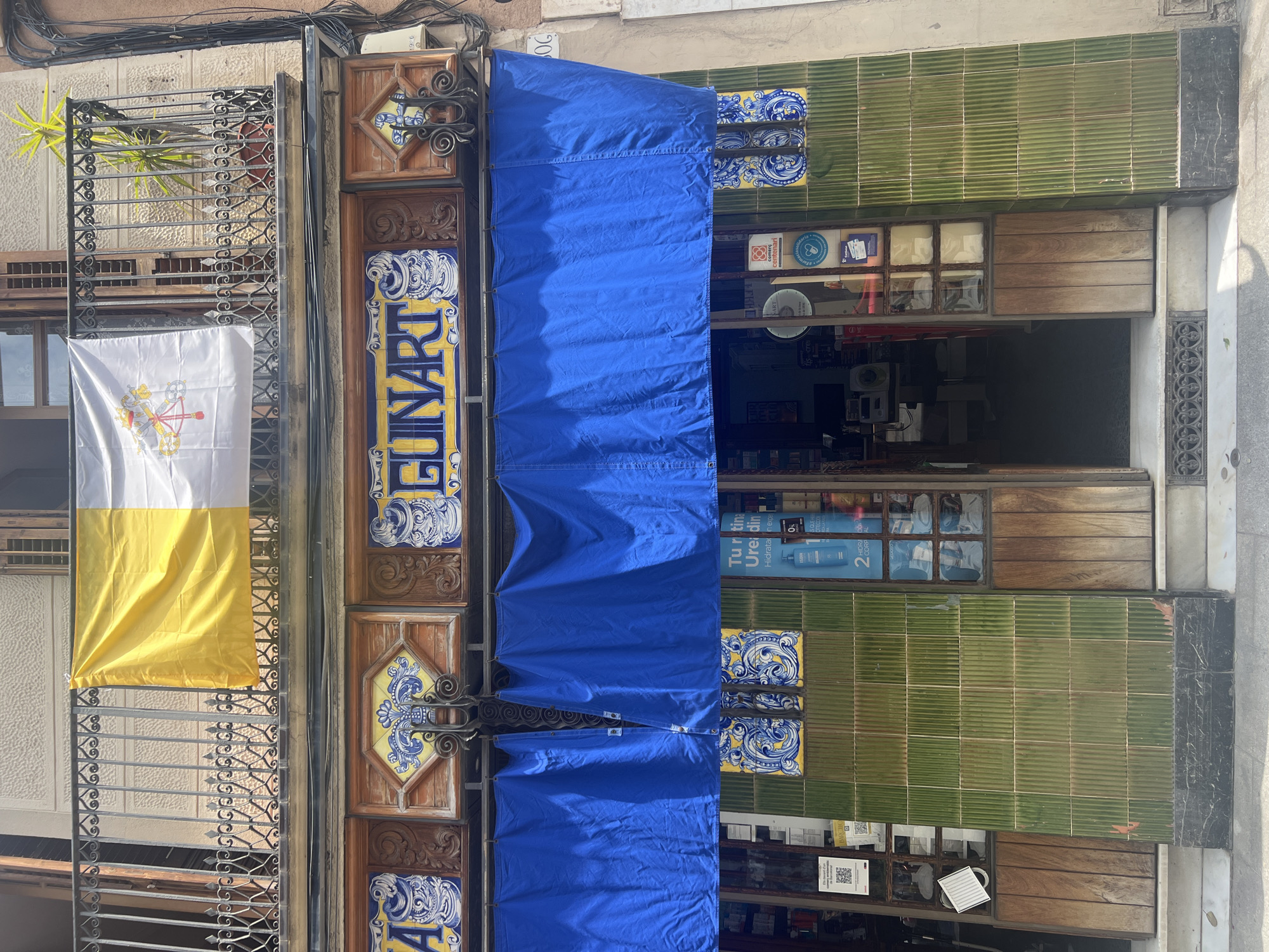 The image shows the exterior of a shop on a street, likely in Barcelona, Spain, due to the architectural style and signage. A Vatican flag, half yellow and half white with the papal insignia, is displayed on the balcony above the shop. The shop's facade features decorative tiles with blue and yellow patterns and wooden accents. A blue awning covers the shop entrance. The shop itself appears to sell a variety of products, as indicated by the shelves and items visible through the glass doors and windows. The street in front of the shop is paved, and there is a doormat at the entrance. The overall impression is of a traditional shop in an urban setting.