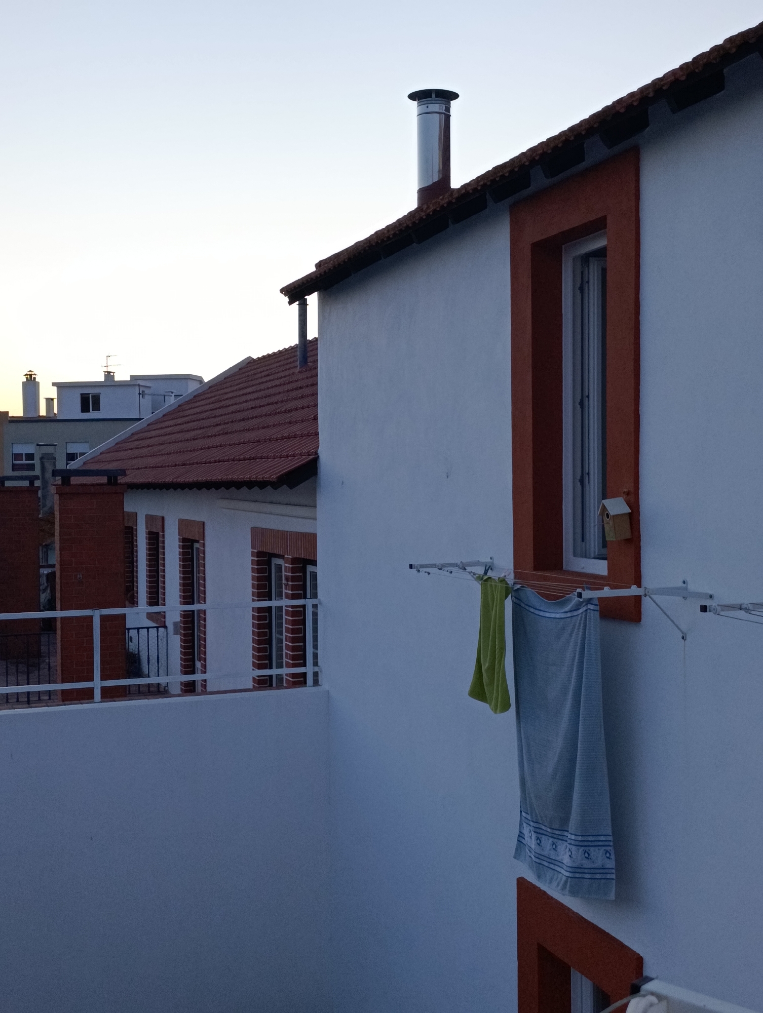 The image shows a view of rooftops and building exteriors in Setúbal, Portugal. In the foreground, a white building with an orange-trimmed window is prominent, with a clothesline holding two towels hanging outside. To the left, another building with a red tile roof and red brick accents is visible, featuring multiple windows and a balcony with a white railing. In the background, more buildings with tiled roofs and various architectural styles can be seen against a pale sky. A chimney is present on the roof of the white building. The scene suggests a residential urban environment.