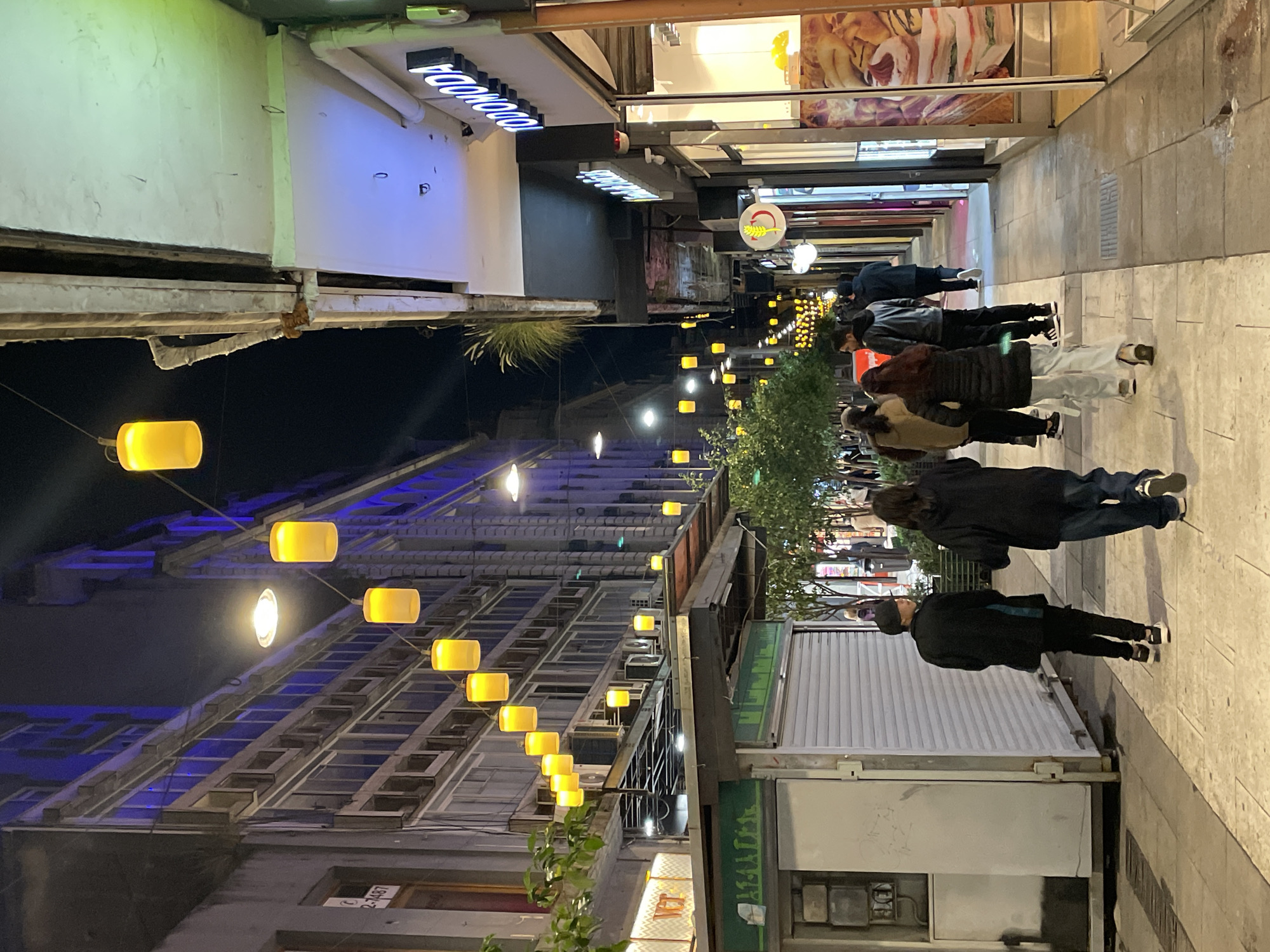 The image captures a street scene in Buenos Aires, Argentina, at night. A line of people stroll along a narrow pedestrian street lined with shops and eateries. Strings of yellow barrel-shaped lights stretch overhead, illuminating the street. The architecture is varied, with buildings ranging from older structures with exposed concrete framework to modern storefronts with bright signage. A sign reading "Foodoo" is visible. People are dressed in casual attire.