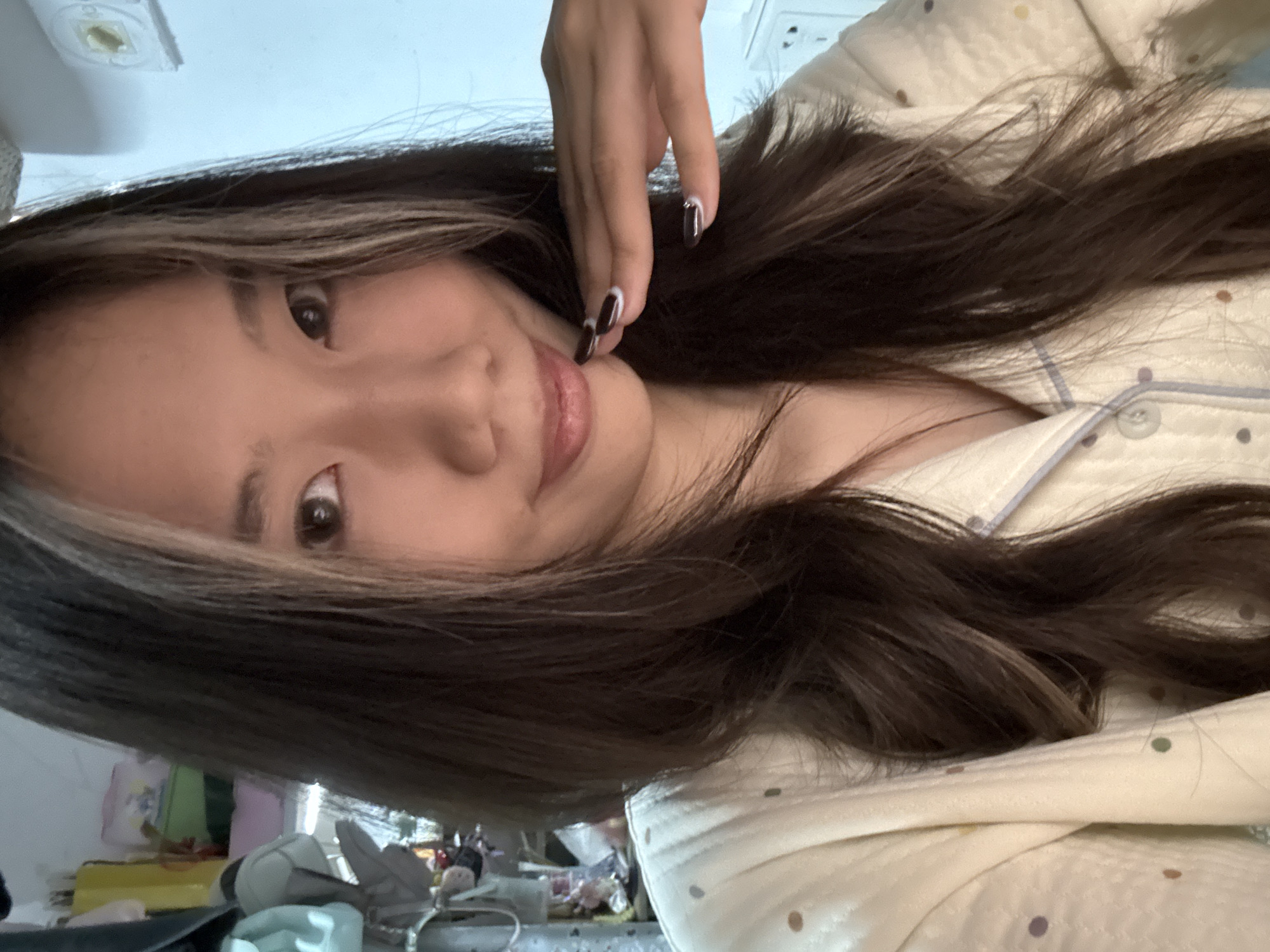The image portrays an East Asian female indoors within a residential setting in Nan'an District, China. She has long, dark brown hair with distinct lighter streaks framing her face and styled in layers. Her dark eyes are directed toward the camera, and she has a subtle smile. She wears a cream-colored long-sleeved top, consistent with pajamas, featuring small, multi-colored polka dots and a light blue trimmed collar and button placket. Her right hand is raised, with fingers gently curled, resting against her lower chin. Her fingernails are painted a dark color with white tips. The background to the left side reveals a cluttered area with various items, including light blue, gray, and pink bags, along with boxes on shelving. The background to the right consists of a light blue wall featuring a white electrical outlet.