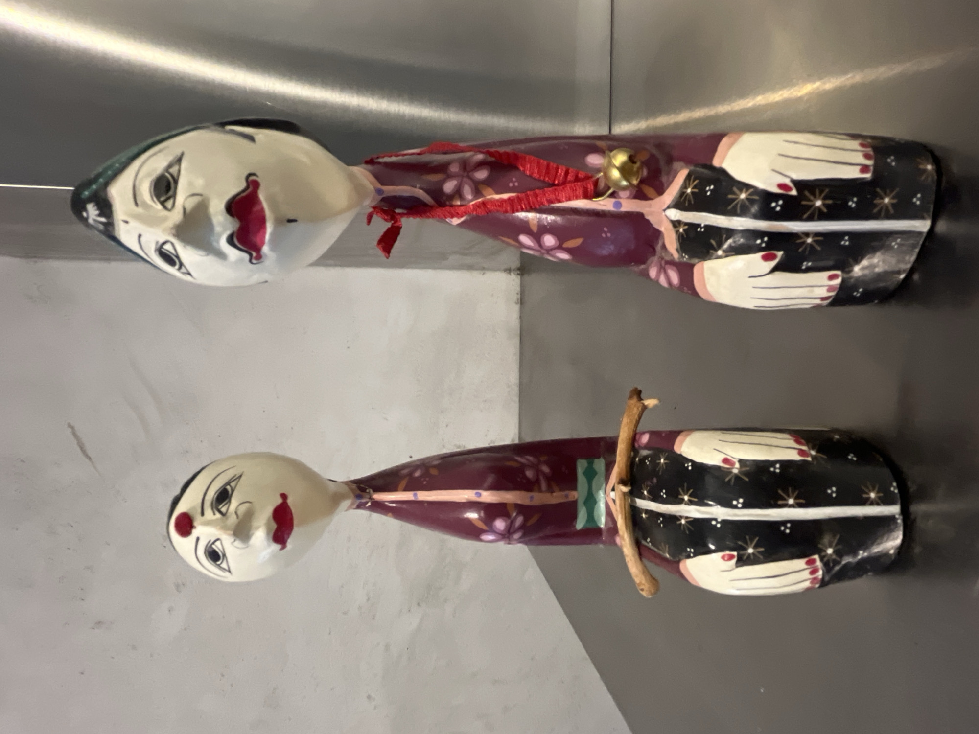 Two hand-painted wooden dolls lie side-by-side. The dolls have white faces with painted features, including black hair, red lips, and distinct eyes. They are dressed in reddish-purple robes adorned with floral patterns. The bottom of each doll is painted black with white and gold star-like designs and white hand-like shapes. The upper doll has a red ribbon around its neck with a golden bell. The lower doll has a thin, light brown twig or small branch attached to its body. The background shows a shiny, light-colored surface, possibly metal, and a neutral-toned wall. There is no indication of Budva, Montenegro in the image.