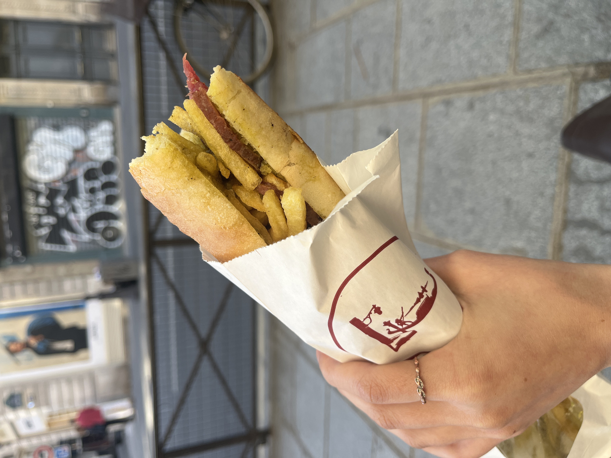 A close-up, eye-level shot shows a person's right hand holding a large, open-faced baguette sandwich. The baguette is split lengthwise and generously filled with golden-brown French fries and thin slices of reddish cooked meat or sausage, some pieces peeking out from the top. The sandwich is wrapped in white paper, which features a distinctive red logo on its side. The logo depicts a stylized silhouette of a person, possibly a vendor, serving food from a stall or counter to another figure. A delicate, silver-toned ring is visible on the ring finger of the hand holding the sandwich.
The background is blurred, suggesting an outdoor, urban setting in Paris, France. To the left, a textured wall or building facade with some light-colored, abstract graffiti or signage is faintly visible, along with part of a bicycle wheel higher up. To the right, a plain stone or concrete wall with horizontal mortar lines provides a muted backdrop. The lighting suggests it is daytime. The scene captures a casual, on-the-go street food experience.