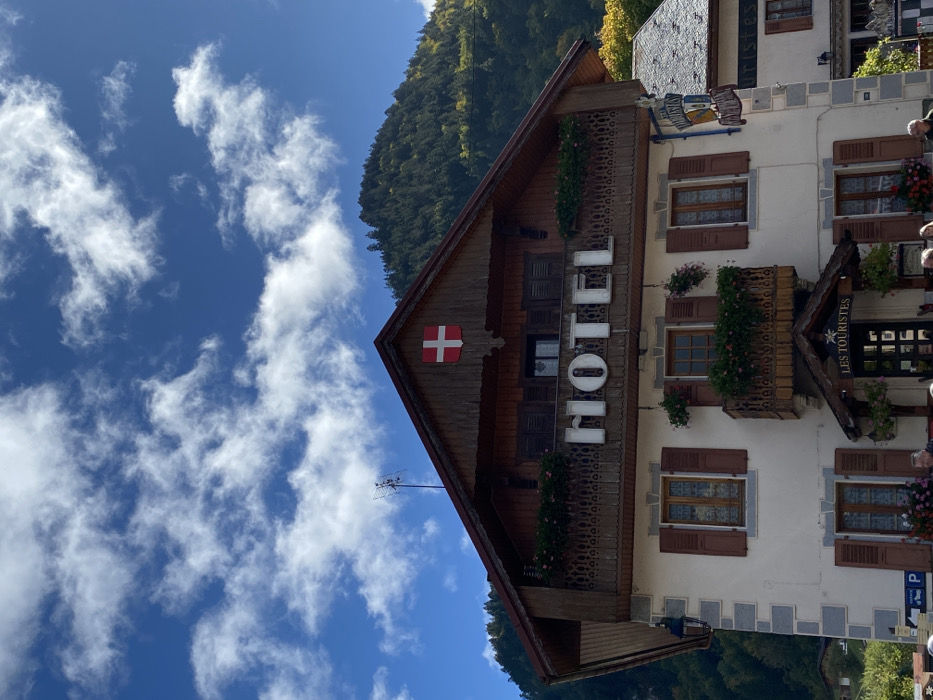 This image displays the Hotel Les Touristes, located in Abondance, France, under a bright blue sky with scattered white cumulus clouds. The building exhibits a traditional Alpine architectural style, featuring a prominent dark brown wooden gabled roof with overhangs. A red shield with a white Swiss cross is affixed to the wooden gable, and a television antenna protrudes from the roof peak.
The upper section of the hotel's facade is constructed of dark brown wood, featuring a balcony with intricately carved wooden railings and window boxes containing pink flowers. Large white dimensional letters spelling "HOTEL" are mounted on this balcony's railing. Below, the building transitions to light-colored (off-white) rendered walls, accentuated by grey stone quoins at the corners. Multiple windows with brown wooden shutters are visible across these lower floors.
The main entrance is positioned beneath a small, covered awning, above which a smaller balcony adorned with flowers is situated. A sign bearing the name "LES TOURISTES" is displayed above this entrance. Additional hanging flower baskets are suspended from balconies and window areas. A black metal lantern is affixed to the left corner of the building's facade. Partially visible at the bottom of the frame are blurred figures of individuals and portions of parking signage. In the background, steep, forested mountains rise, displaying a mix of green and some yellow foliage, indicative of late summer or early autumn.