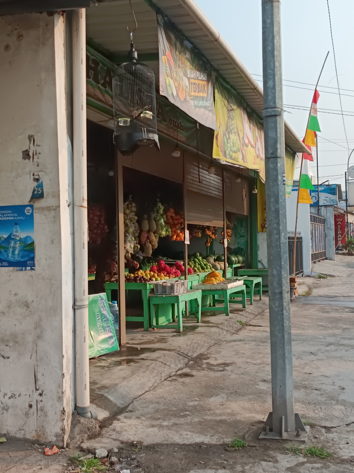A fruit stand is set up on the side of a street in Sewon, Indonesia. The stand has a green roof and displays a variety of fruits including dragon fruit, watermelon, mango, and bananas. A birdcage hangs from the roof of the stand. There are several flags on poles in front of a building to the right of the fruit stand. The ground in front of the stand is paved.
