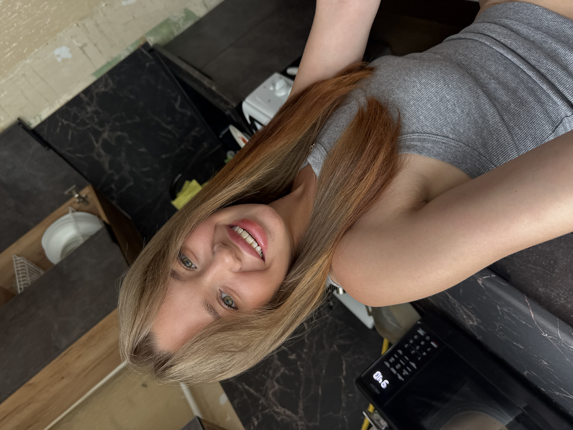 In a dimly lit kitchen that appears to be undergoing renovation, a young woman with blonde hair and green eyes smiles at the camera. She is wearing a grey ribbed tank top and her hair is partially covering her left shoulder. In the background, dark cabinets with a marble-like pattern are visible, along with exposed plaster walls. To the left, a black microwave with a digital display showing "9:40" is present. Other kitchen appliances and shelves can be seen in the background, suggesting a functional yet incomplete living space. The overall scene is a selfie taken in a domestic environment.