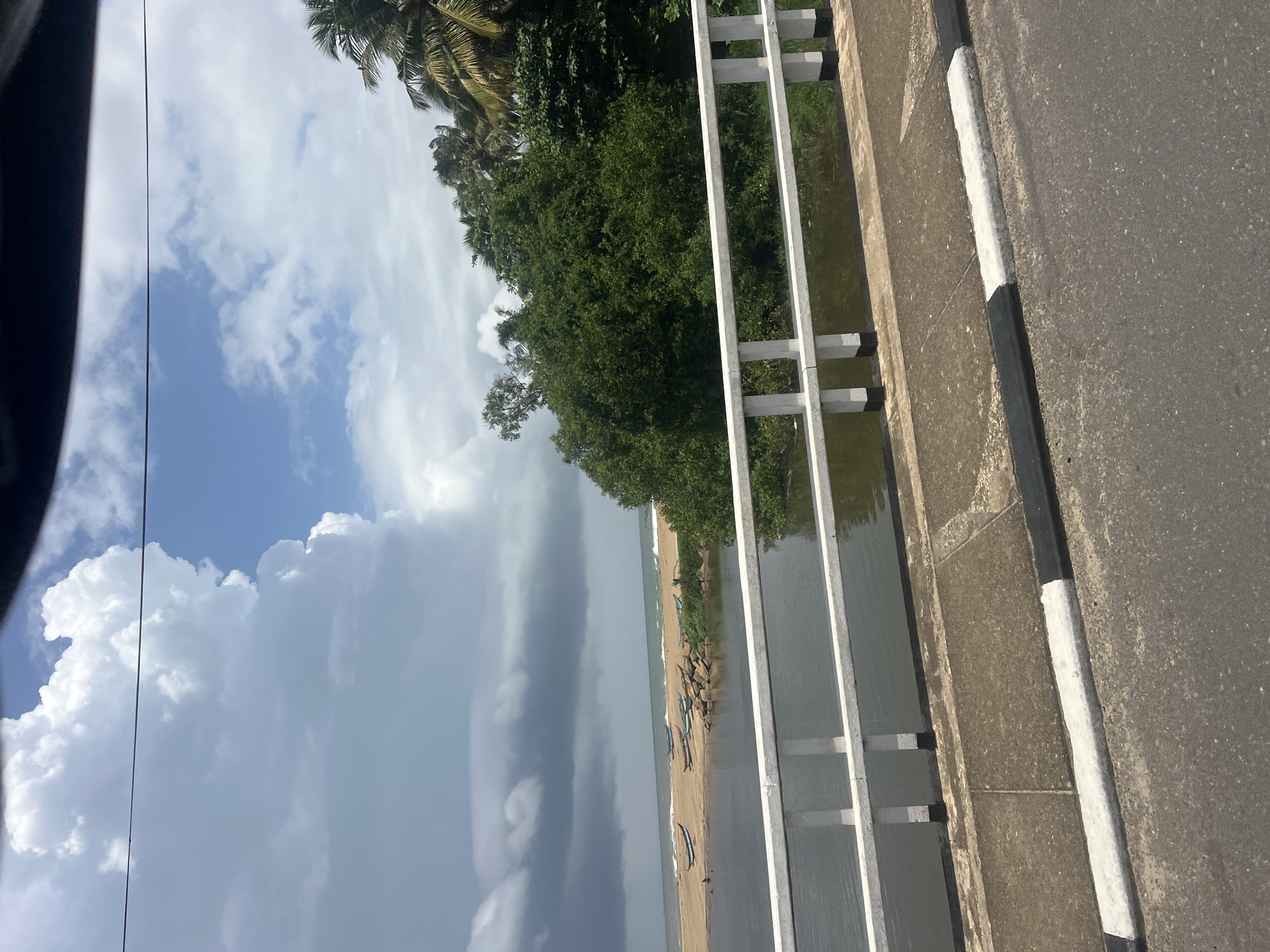 The image depicts a scenic view from a bridge in Rajgama, Sri Lanka. The foreground features a concrete bridge with a white railing overlooking a body of water. The water reflects the sky and surrounding greenery. Beyond the water, a sandy beach is visible, with several small boats resting on the shore. The ocean stretches out in the background, meeting a sky filled with dramatic clouds, some of which appear dark and ominous, suggesting an approaching storm. Lush green trees and palm trees line the coastline, adding to the tropical ambiance. The overall scene captures a moment of natural beauty and tranquility, with a hint of impending weather change.
