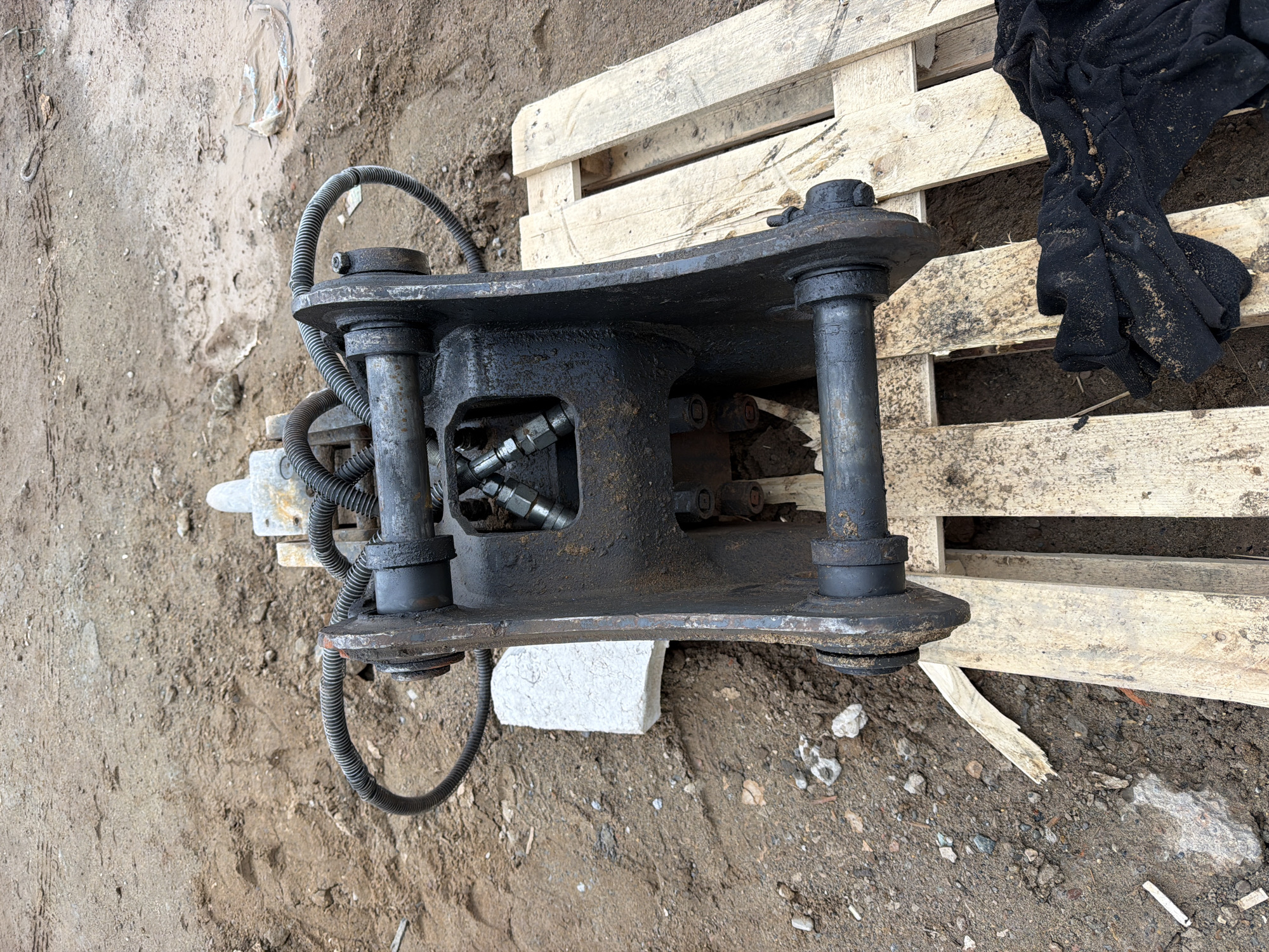 The image shows a hydraulic jack attachment on the ground, likely in an industrial area. The jack is black and appears to be used. It sits partially on a wooden pallet. The ground is dirt and gravel. A white brick is partially supporting the jack. The location is possibly in the city of Chaikovsky, Russia (based on the user's request for the location). There are no people visible in the image. A piece of black cloth is on the pallet. The scene seems to depict construction or maintenance.
