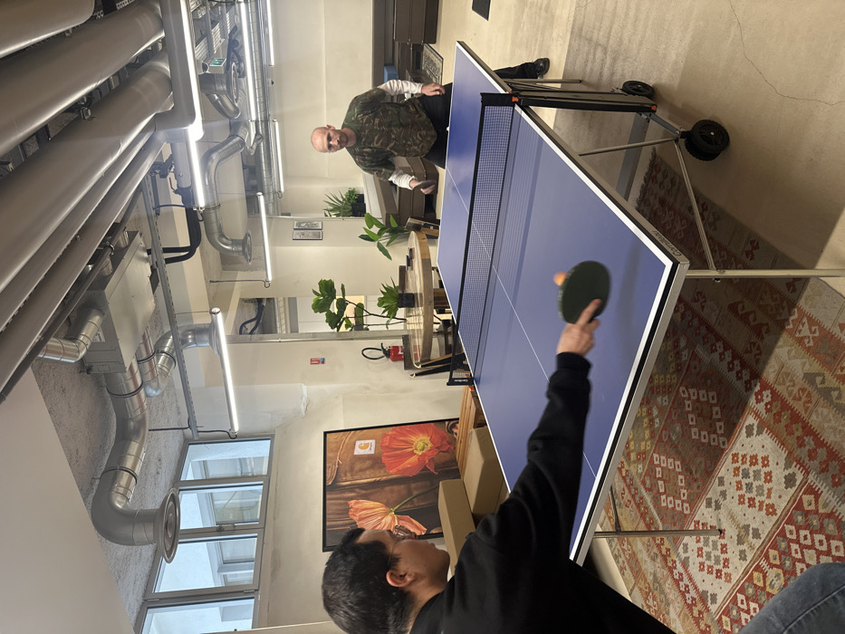 The scene depicts two adult males engaged in a game of table tennis within a contemporary industrial-style interior. The specific location is identified as the Carhartt WIP office at 4 Rue des Minimes, 75003 Paris, France.
In the foreground, a male with dark hair and glasses, wearing a black long-sleeved shirt, is positioned on the left, viewed from the rear-left. He is actively striking an orange ping-pong ball with a dark green paddle, his arm blurred by motion. He stands on a patterned kilim-style rug.
Opposite him, on the right side of the blue Cornilleau table tennis table, stands a bald male with a beard, wearing a camouflage-patterned t-shirt layered over a white long-sleeved shirt and dark trousers. He holds a paddle, poised to play. This player stands on a light-colored polished concrete floor.
The room's ceiling is concrete with exposed metallic ventilation ducts and linear strip lighting fixtures. Large windows are visible on the far left. In the mid-ground, a large framed artwork featuring orange poppies and a 'Carhartt' logo rests above stacked items, possibly cardboard boxes. Other background elements include round wooden tables with chairs, multiple potted plants, a red fire extinguisher, and dark grey modular seating units. A "no smoking" symbol is affixed to a pillar, and a green exit sign is visible on the ceiling.