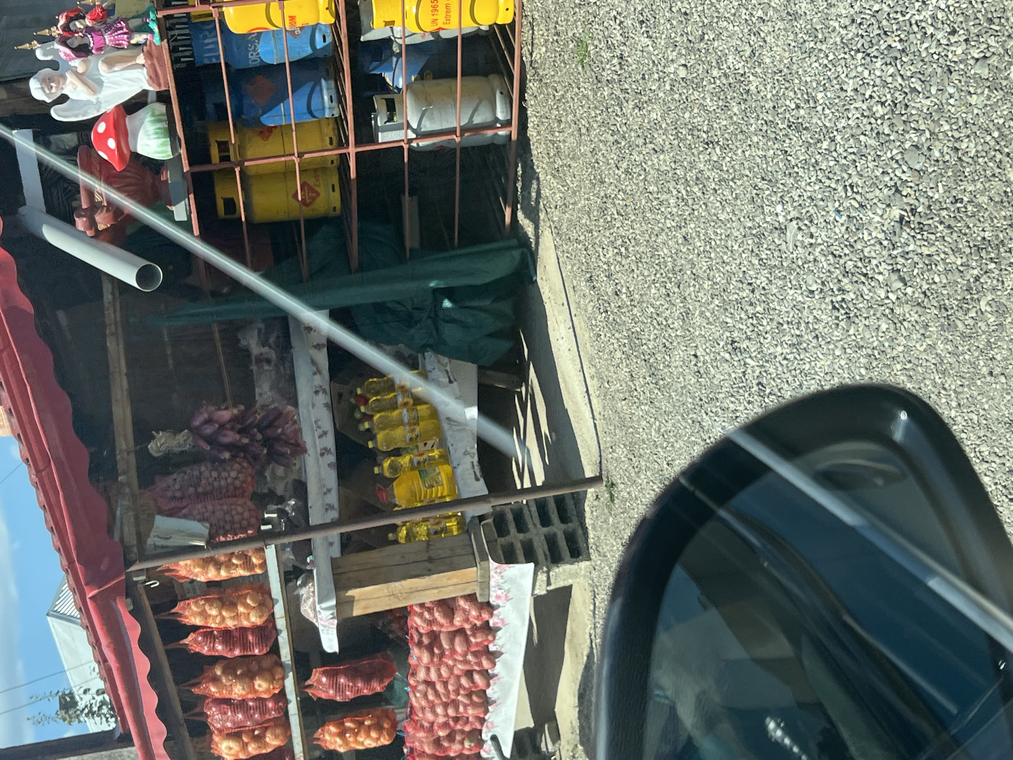 In Băișești, Romania, a roadside stall displays a variety of goods. On the left, nets of onions and red onions hang from the stall's awning. Below these, bags of potatoes are arranged on a table covered with a patterned cloth. In the center, bottles of yellow liquid, likely cooking oil, are lined up. To the right, shelves hold propane gas cylinders in various colors, including yellow, blue, and gray. A few decorative items, such as a ceramic mushroom, an angel figurine, and two dolls, are also visible on the shelves. The ground in front of the stall is covered in gravel, and a portion of a car's windshield and side mirror is reflected in the foreground. The sky above is blue with some clouds.