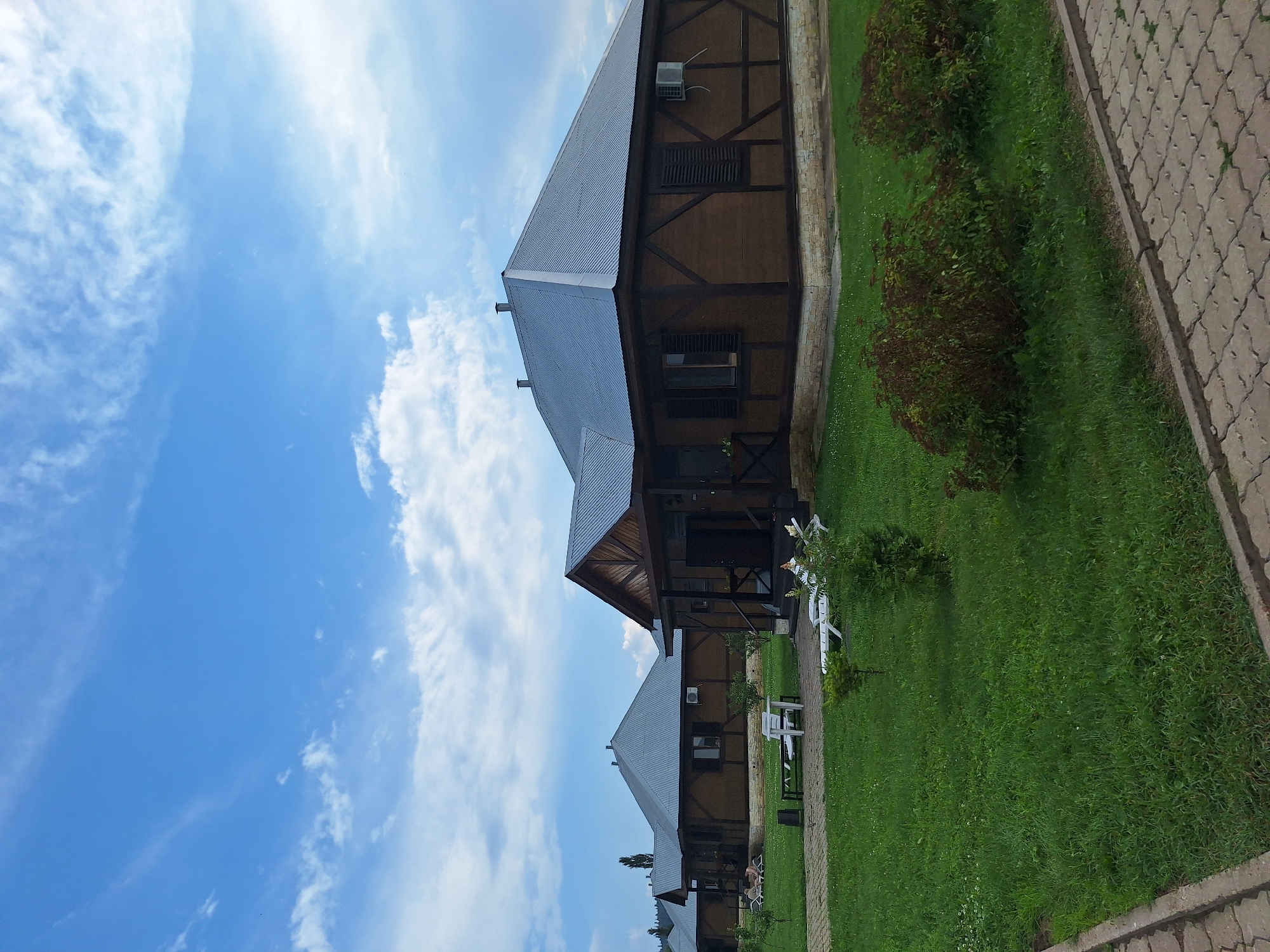 In Atamanovka, Russia, a resort complex features several identical, rustic wooden cabins with dark metal roofs. The cabins are arranged in a row, with the nearest one to the right featuring a porch with dark wooden railings and a dark door. The cabins have wooden siding with dark timber framing accents and stone bases. Each cabin is equipped with an outdoor air conditioning unit.
The grounds are covered in lush green grass, with a paved walkway made of light grey interlocking stones bordering the area. Several white lounge chairs are scattered on the lawn in front of the cabins, some with umbrellas. Small, manicured bushes and trees are strategically placed throughout the landscape. In the distance, more cabins are visible, as well as a treeline and hills under a bright blue sky with scattered white clouds. The atmosphere appears calm and serene, suggesting a leisurely retreat.
