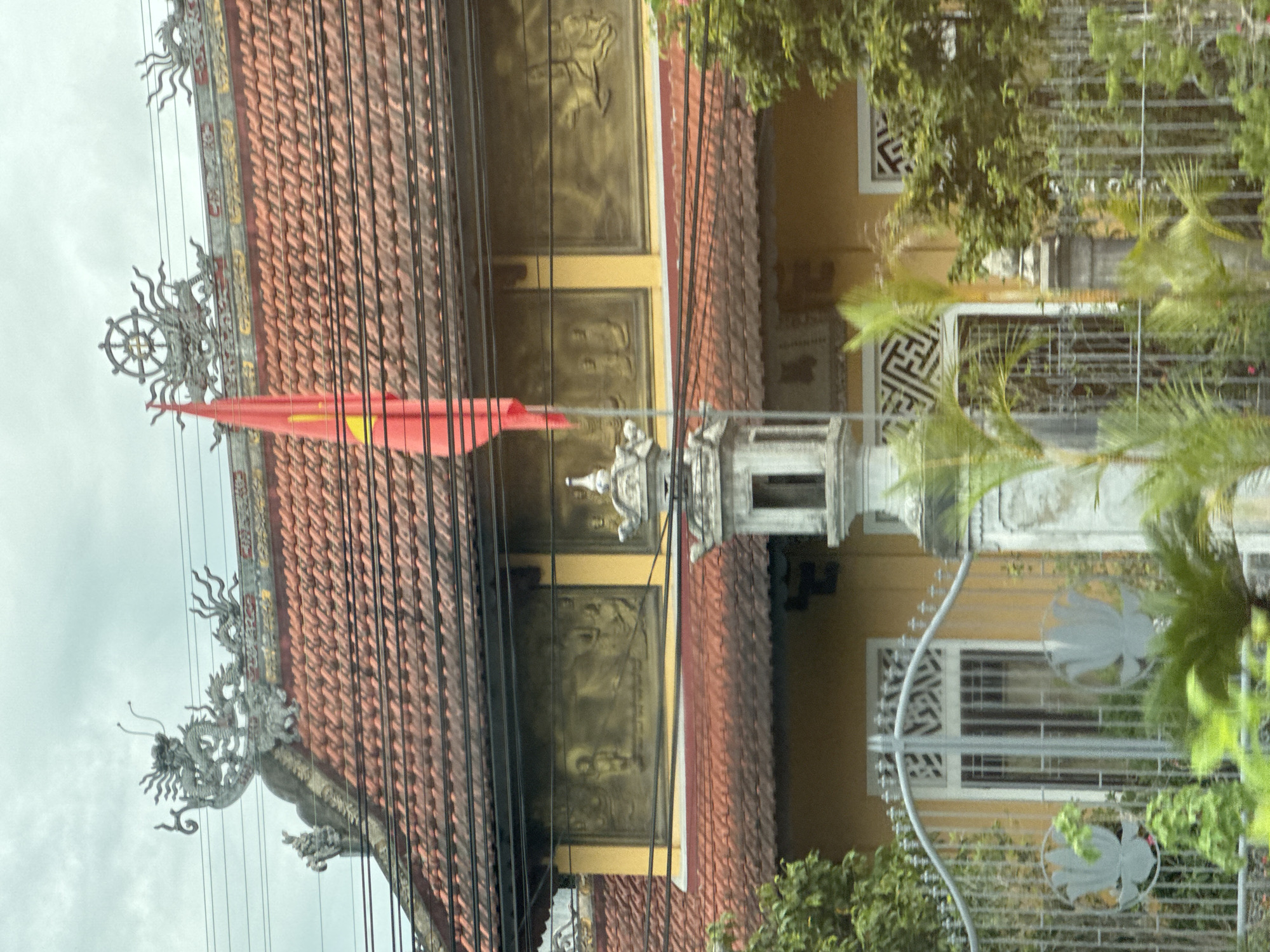 The image shows a building with a red tiled roof, likely a temple or pagoda, in Kim Long Ward, Vietnam. A red flag with a yellow star, the flag of Vietnam, is prominently displayed, attached to a flagpole. The roof is adorned with decorative ceramic figures, including a dragon on the left and what appears to be a Dharma wheel on the right. Below the roofline, there are ornate gold-colored relief panels depicting figures in what seems to be a religious or historical context. The building's exterior walls are painted yellow. In the foreground, a decorative metal gate with lotus motifs encloses the property. A white stone structure, possibly a lantern or a small shrine, stands near the gate. Lush green foliage, including palm trees, surrounds the building and the gate, suggesting a tropical or subtropical environment. Power lines run horizontally across the upper portion of the image. The sky is overcast.