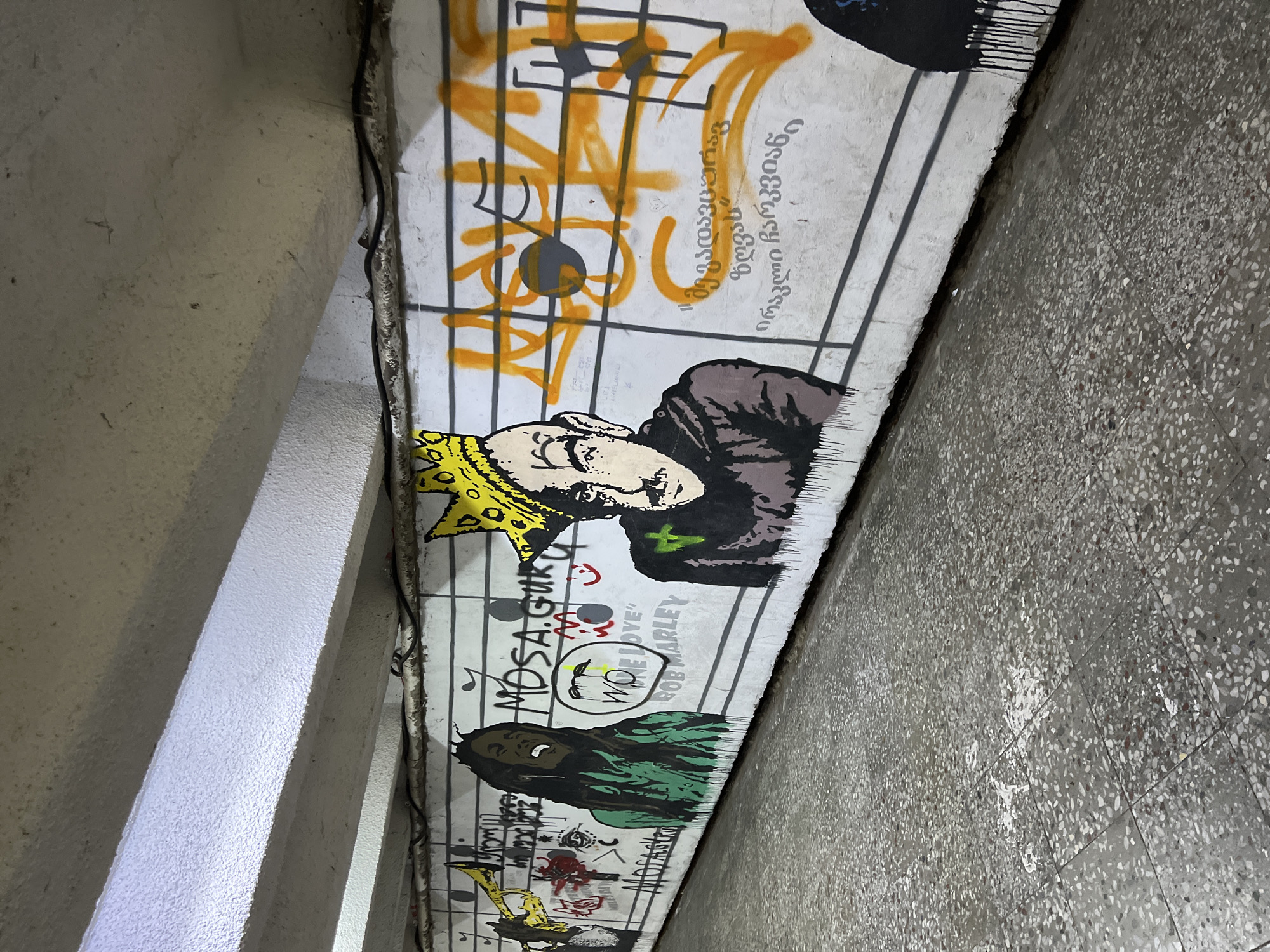 The image captures graffiti art on a concrete wall within an underpass or similar structure in Tbilisi, Georgia. The artwork features a series of figures and musical notations against a white background with painted lines resembling a musical staff.
Prominent figures include portraits of individuals, one wearing a yellow crown and another appearing to be Bob Marley. Inscriptions such as "MOSA.GURU" and "WOKE LOVE" are visible near the figures. There are additional abstract and symbolic elements, including musical notes and what appear to be stylized letters in an unknown language. The color palette primarily consists of yellow, orange, black, and green accents over the white background.
The setting is characterized by a concrete structure with visible seams and edges, along with adjacent concrete elements and an overhead electrical conduit. The floor appears to be a tiled surface with a speckled pattern.