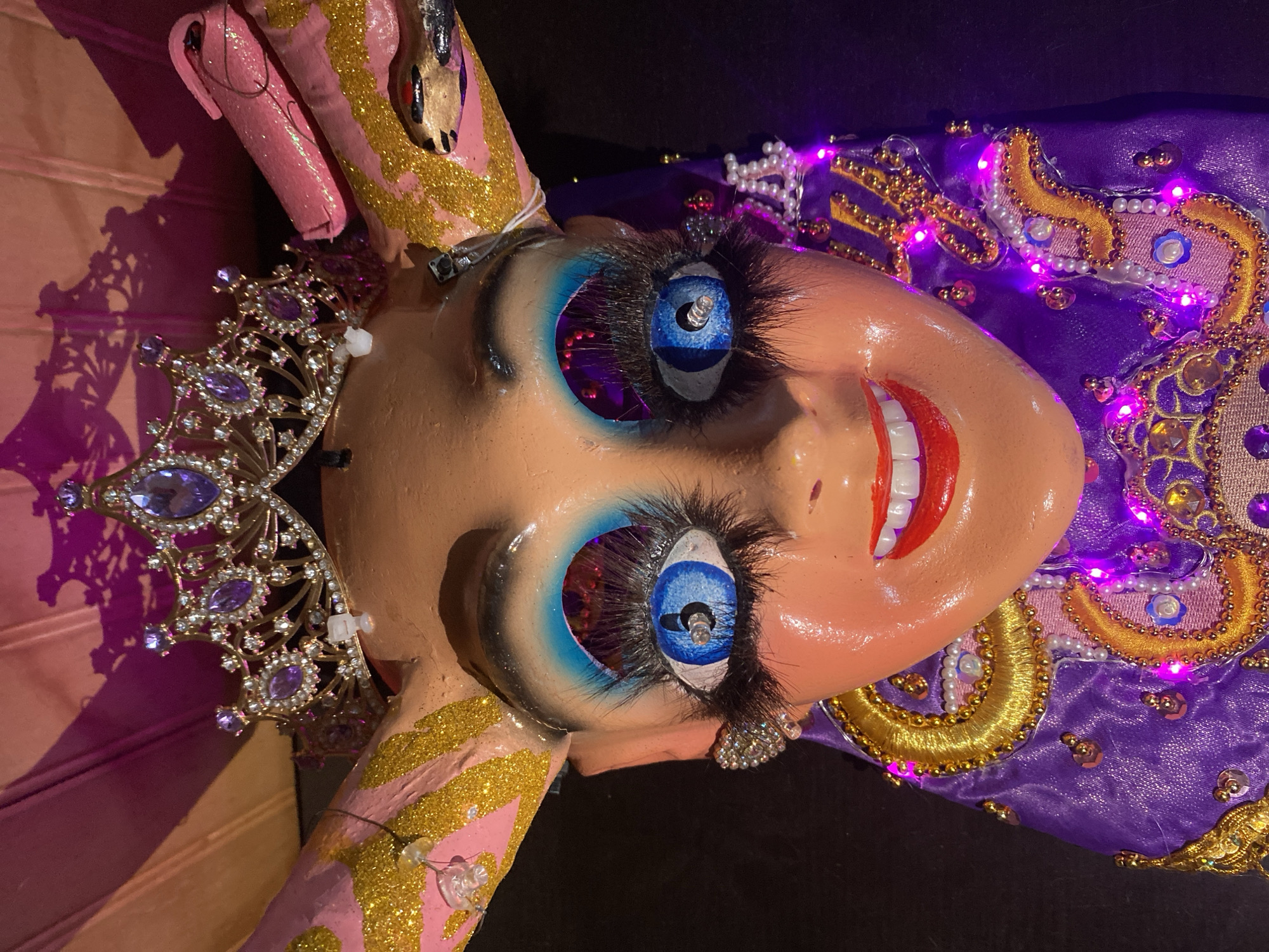 The image shows a close-up, rotated view of an elaborate, possibly handcrafted doll or mannequin head. The head is pale with exaggerated features: large blue eyes with long eyelashes, bright red lips showing teeth, and heavily applied blue eyeshadow. The head is adorned with a jeweled crown on the left side and is partially covered with purple fabric embroidered with gold details and sequins, interspersed with small purple lights. The object’s right arm is visible, painted pink with gold stripes, and topped with a snake figure. The lighting is low. The location in the city of Arica, Chile is not discernible from the visual information provided.