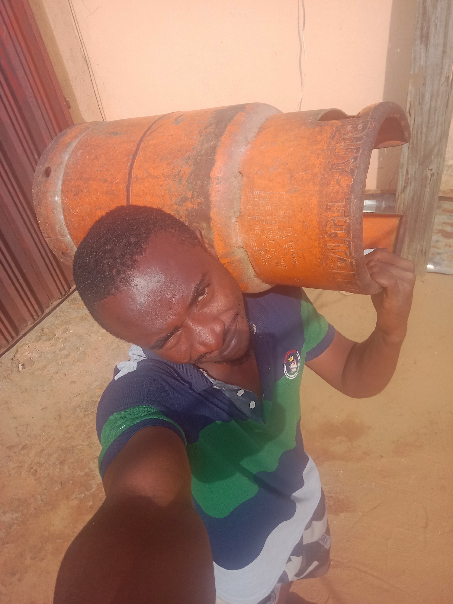 A man stands outdoors, carrying an orange, cylindrical gas tank on his left shoulder. The tank is labeled "TOTAL" and has other printed text, including "BUTANE NET 12.5KG." The man is facing the camera and appears to be taking a selfie. He is wearing a blue and green polo shirt and patterned shorts. The background consists of a peach-colored wall, a brown door or paneling, and a wooden post. Based on the OCR text on the tank mentioning "Nigeria," and per information you requested, the location is Obosi, Nigeria.