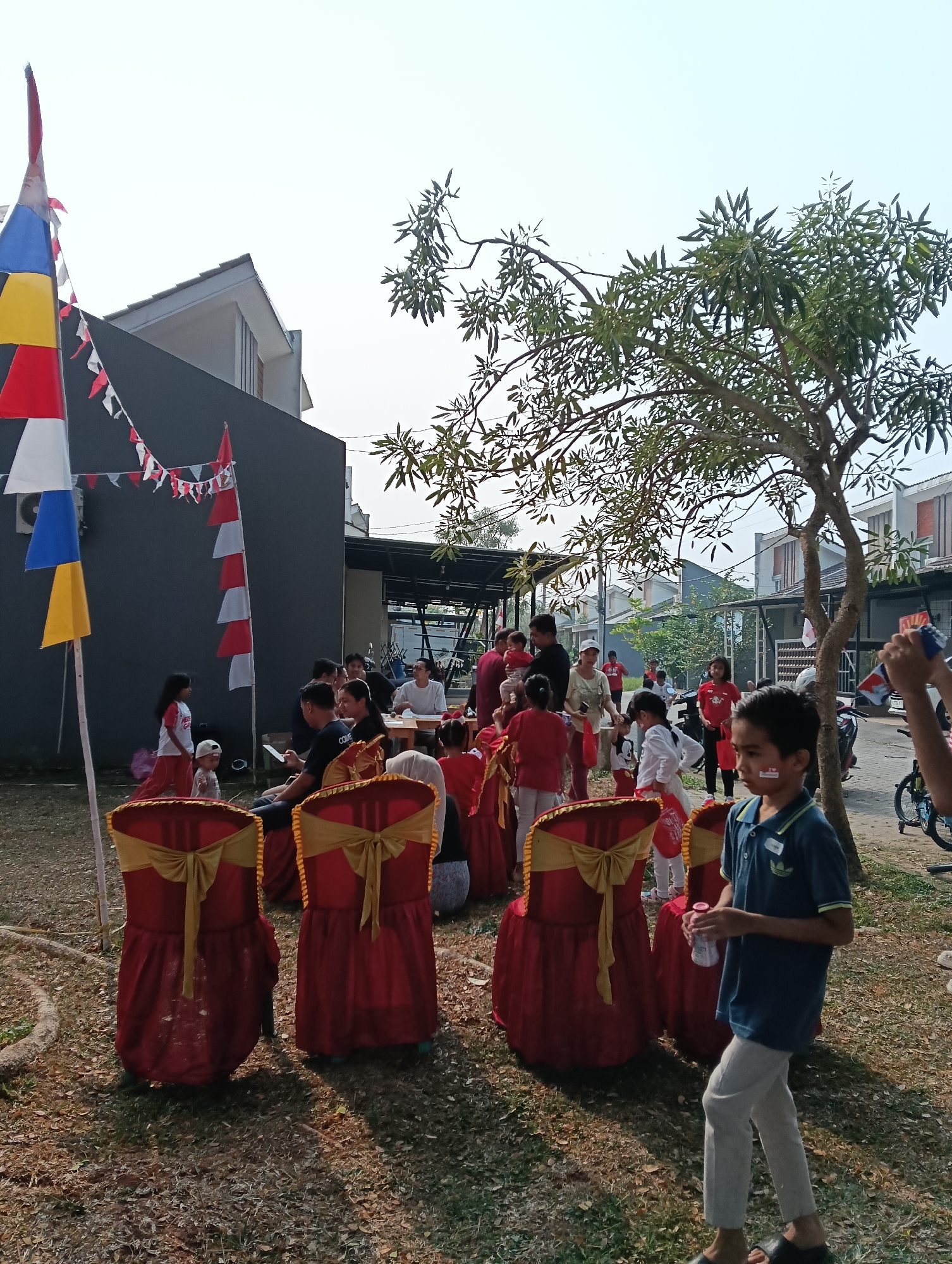 In Parung Panjang, Indonesia, under a clear sky on a sunny day, an outdoor gathering is underway. The scene is decorated with Indonesian flags, featuring alternating red and white triangular banners strung between poles and along the side of a dark gray building. A tall flag pole with red, yellow, and blue triangular flags also stands prominently. The ground is covered with dry leaves and patchy grass, with a concrete border visible in the foreground.
Numerous people are present, with many seated on chairs draped in red fabric and tied with golden sashes. These chairs are arranged in rows. A group of children and adults are standing and interacting. Some children are wearing red shirts, while others are in casual attire. A young boy in a blue polo shirt and light-colored pants walks towards the right of the frame, holding a pink bottle. Another child in the foreground has a red and white stripe painted on his cheek. In the background, modern houses with gray roofs are visible. A tree with sparse green leaves provides some shade. A bicycle is parked to the right, and a motorcycle is visible behind a group of people. The overall atmosphere suggests a festive community event.
