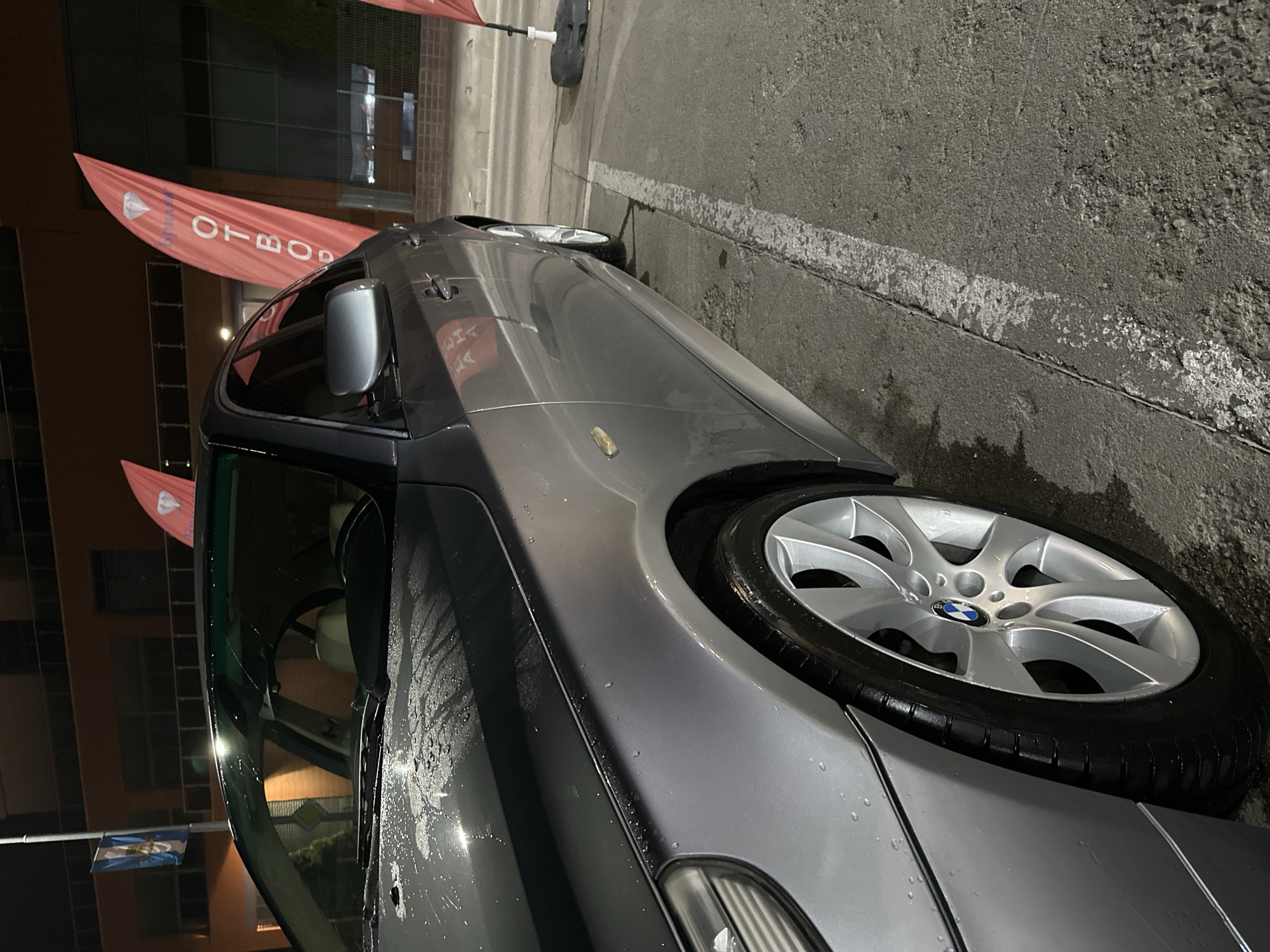 The image shows a gray BMW car parked on an asphalt surface, possibly a street or parking lot, next to a painted white line. In the background, there are buildings and flags. The car appears to be wet, suggesting recent rain or washing. Based on the context of Plovdiv, Bulgaria, it is difficult to pinpoint the exact location within the city without more specific landmarks or surroundings.