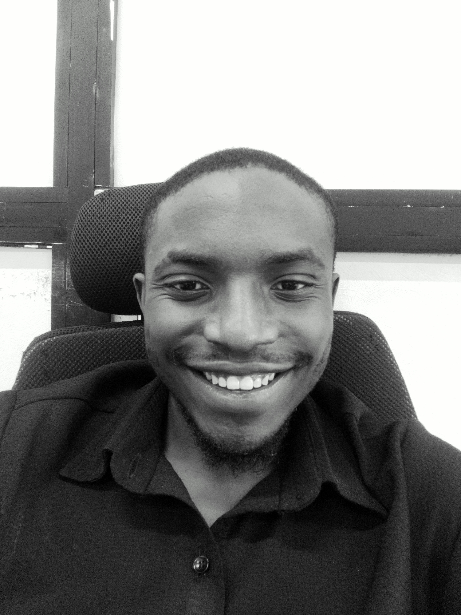 A black and white, medium shot selfie captures a man from the chest up, sitting in an office chair. He is smiling broadly, revealing his teeth. The man has short hair and a goatee. He is wearing a collared, dark-colored shirt. Behind him, the headrest of the chair is visible, along with a dark wooden partition and a white wall. The scene appears to be an indoor setting, possibly an office in Abuja, Nigeria.
