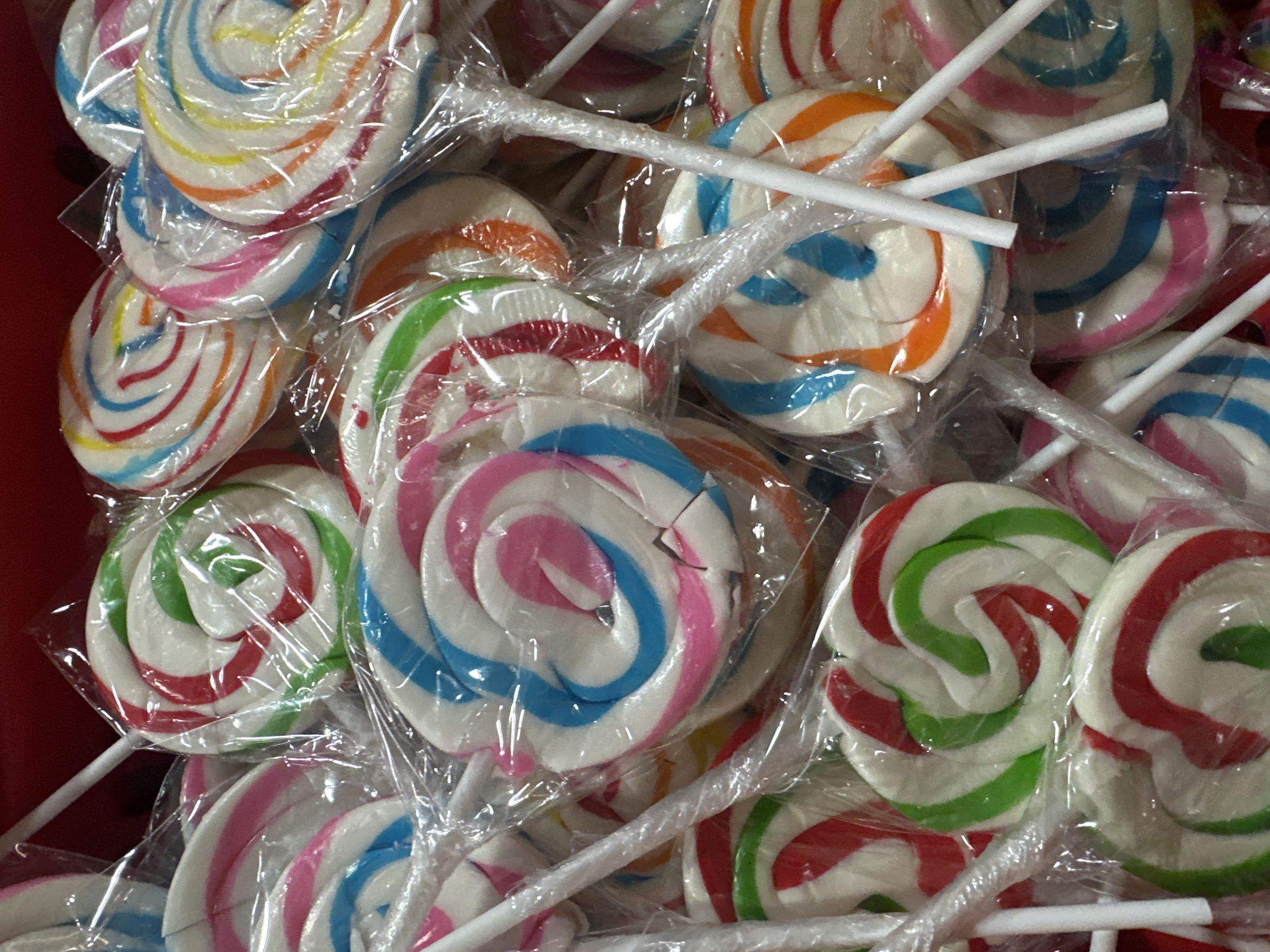 A collection of individually wrapped, swirl lollipops with white sticks is seen. The lollipops are of various colors, including combinations of white with blue, pink, red, green, orange, and yellow. Some wrappers appear damaged. The background is a reddish color.