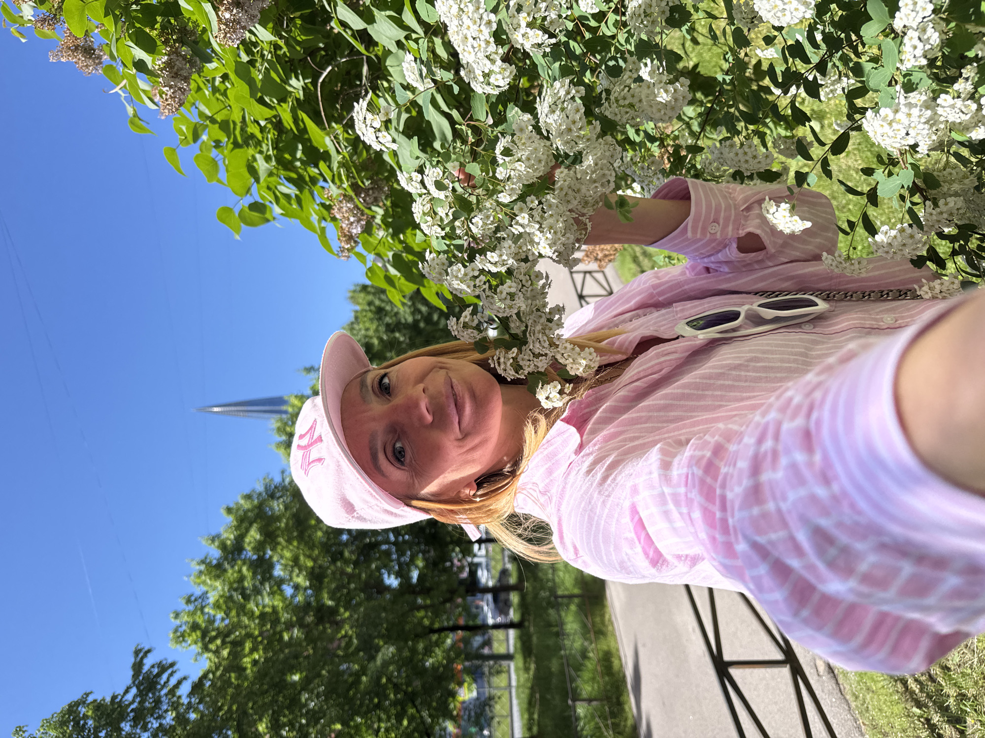 An outdoor selfie captures a woman among flowering bushes on a bright, sunny day in what appears to be Saint Petersburg, Russia. The image is primarily oriented vertically, showing a clear blue sky in the background.
The main subject is a woman, likely in her 30s or 40s, with blonde hair and blue eyes, smiling gently at the camera. She is wearing a pink and white striped long-sleeved shirt and a pink baseball cap that features a stylized white "M" or similar letter on the front. White sunglasses are clipped to the collar of her shirt.
She is surrounded by and partially obscured by vibrant green foliage and numerous clusters of small white flowers, characteristic of a flowering shrub like Spiraea. These blossoms are prominent in the upper and central parts of the frame, some extending towards her. The setting appears to be a park or a green public space, with a paved path or road and more trees visible in the background, implying a pleasant spring or summer day. A distinct, tall, dark spire or monument rises sharply against the clear blue sky in the mid-ground, a notable architectural feature often seen in Saint Petersburg. The overall atmosphere conveys a sense of contentment and enjoyment of the natural environment.