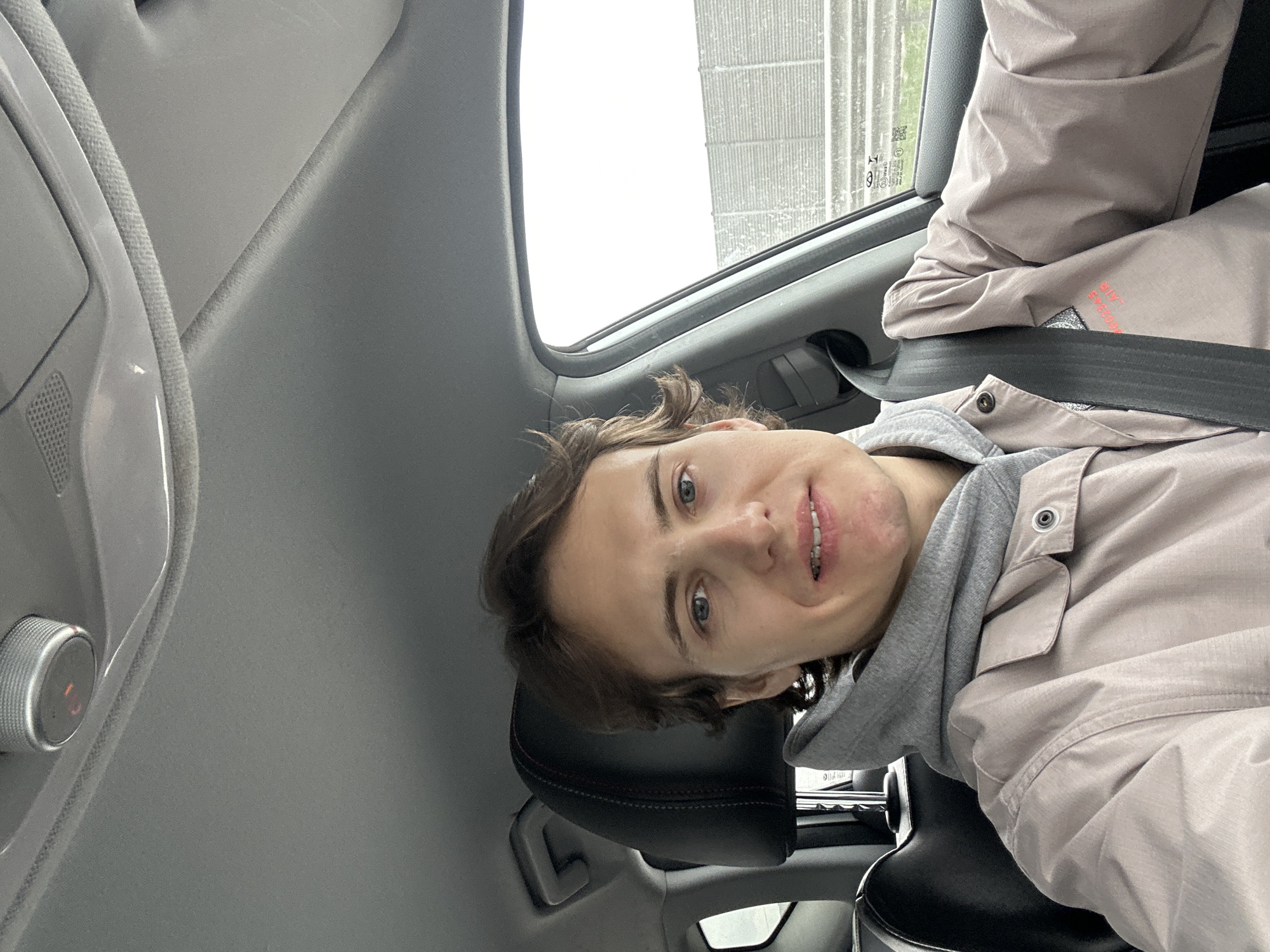 A young individual with short brown hair and blue eyes is seated in the passenger seat of a vehicle. The person is wearing a gray hoodie underneath a beige button-up jacket and a seatbelt. The interior of the vehicle, including the headrest, dashboard elements, and the window showing external weather conditions. The location in Zaozerye, Russia cannot be determined.