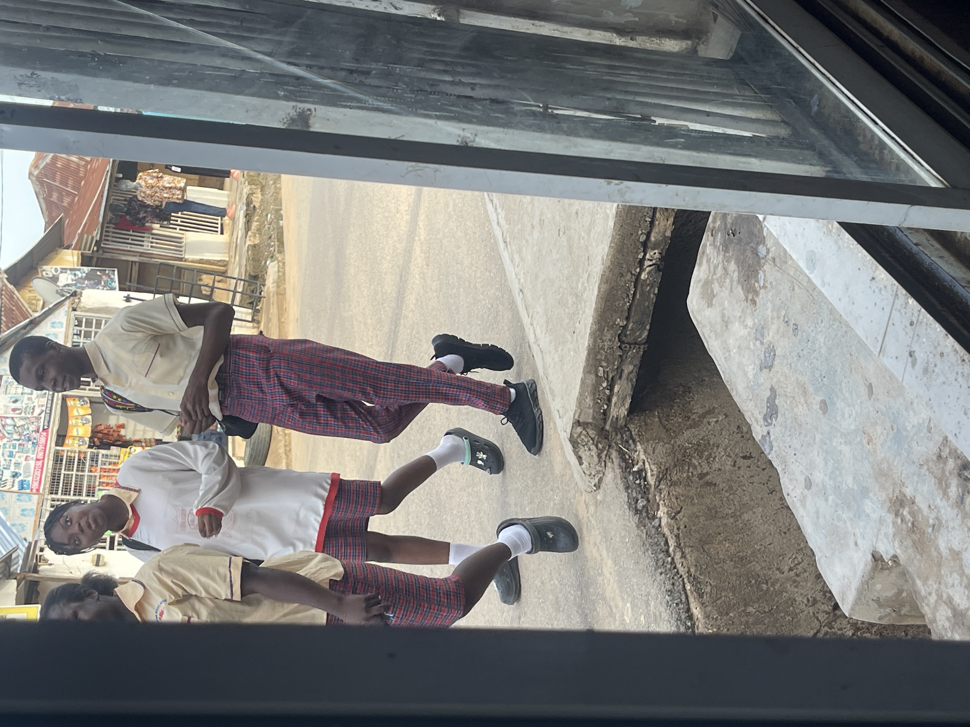 The image depicts a daylight street scene in Abuja, Nigeria, viewed through a window or door frame that partially obstructs the left and right edges. Four individuals, appearing to be young adults or teenagers, walk on a concrete sidewalk from left to right. All are attired in what resembles a uniform: light yellow short-sleeved collared shirts with red trim on the collar and sleeves, paired with red, white, and dark-colored plaid bottoms.
From left to right:
1. A partially visible individual is seen wearing a yellow collared shirt and plaid shorts/skirt. Their feet are clad in dark Crocs with white socks.
2. A female, positioned centrally, looks towards the camera with a slightly open-mouthed expression. She wears a loose white long-sleeved top with red collar and cuff trim over a yellow collared shirt, and plaid shorts/skirt. She holds a clear plastic cup in her left hand. Her feet have dark Crocs adorned with a light green charm, worn over white socks.
3. A male figure, smiling slightly and looking to his right, wears a yellow collared shirt and full-length plaid trousers. He carries a dark backpack on his right shoulder. Black athletic sneakers with white socks are on his feet.
4. A fourth individual, partially visible on the far right, also wears a yellow collared shirt and plaid trousers.
The immediate foreground features a dark vertical structural element. Below the pedestrians, a concrete sidewalk borders an asphalt road, separated by a deep, open concrete gutter or trench. The background comprises a row of single-story commercial buildings across the street, characterized by reddish-brown corrugated metal roofs, some showing signs of rust. These buildings include storefronts, with one displaying mannequins dressed in patterned shirts and trousers. Various posters and signs are affixed to the building facades, and a white satellite dish is mounted on one roof. A wooden ladder leans against a building wall to the left of the mannequins.