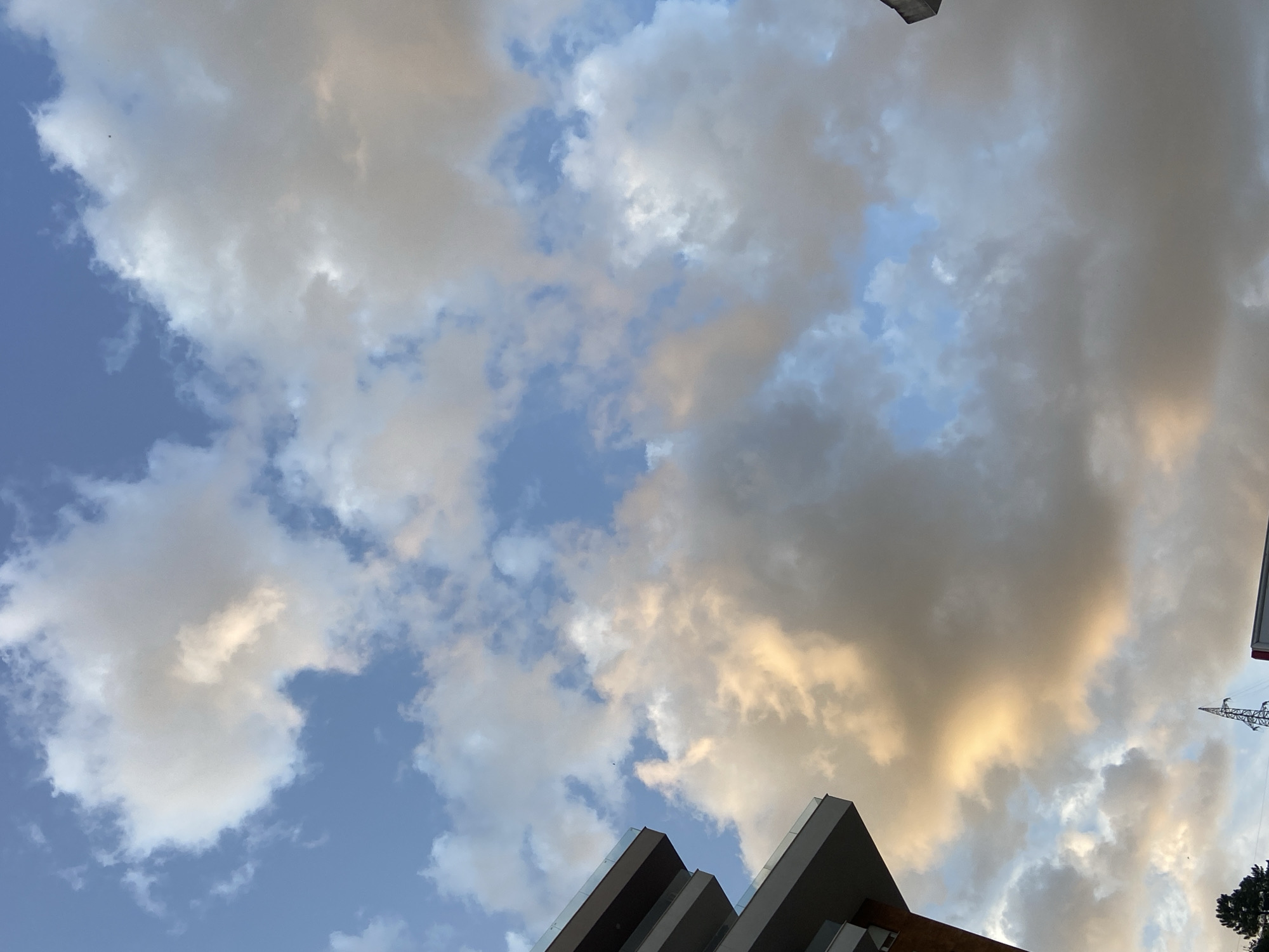 The image shows a view looking up at a sky filled with clouds, with some patches of blue visible. The clouds are a mix of white, grey, and golden hues, likely due to the light of the setting sun. In the lower portion of the image, sections of a modern building can be seen with geometric shapes and reflective surfaces. A power line can be seen at the right edge of the image. The image does not provide conclusive information about the location in Budva, Montenegro.