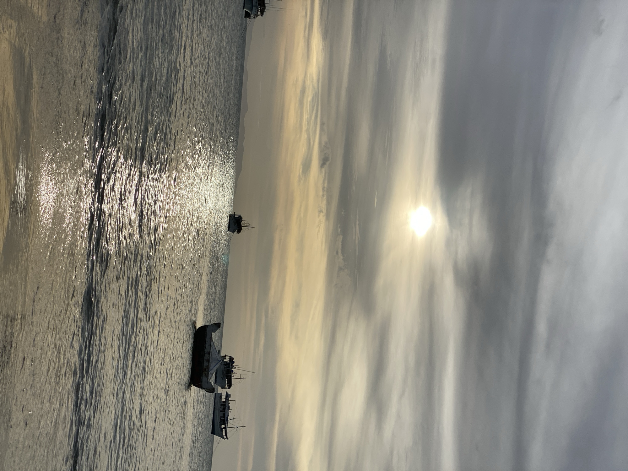 The image depicts a tranquil coastal scene in Juan Griego, Venezuela, at what appears to be late afternoon or early evening. The sky is largely overcast, featuring a mix of grey and yellowish-white stratiform clouds that partially obscure the sun. The sun, a bright, diffuse orb, is positioned in the upper center-right quadrant, casting a prominent shimmering reflection path across the water's surface towards the lower left foreground.
The body of water is calm, displaying gentle ripples. In the immediate foreground, a narrow band of wet sand or very shallow water is visible at the bottom left. Five vessels, consistent with fishing boats, are anchored in the water. A small, dark-hulled boat with a lighter-colored cabin or tarp is positioned in the midground, slightly right of center, appearing aged. Behind it and further out, three larger dark-hulled vessels are visible, equipped with masts and superstructures, appearing as silhouettes against the brighter horizon. A fifth large vessel is partially visible on the far left edge of the frame. No individuals are discernible on the boats or shoreline, and no direct actions or interactions are depicted. Distant landmasses are faintly silhouetted on the horizon line in the background.