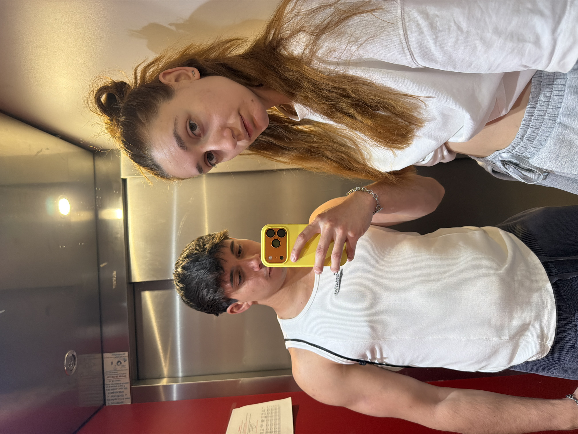 An image captured inside an elevator shows two individuals taking a mirror selfie. The image is oriented sideways, with the top of the subjects' heads pointing to the left.
The individual closer to the top of the frame (when upright) is a woman with long, reddish-brown hair, some of which is tied in a bun while the rest flows freely. She wears a loose white long-sleeved top and grey sweatpants, with her midriff partially visible. A silver ear cuff is visible on her left ear and a silver chain bracelet on her left wrist. She looks directly at the camera with a neutral expression.
The individual closer to the bottom of the frame (when upright) is a man with short, dark hair. He is wearing a white ribbed tank top, possibly with a faint logo on the chest and a black stripe detailing on the shoulder. He holds a bright yellow smartphone in his right hand, which he is using to take the picture, reflecting both subjects in the elevator's polished metallic surface. A silver chain bracelet is visible on his left wrist. His gaze is directed towards the phone screen.
The setting is an elevator interior, featuring reflective metallic walls and a vibrant red floor. Artificial lighting illuminates the space. In the lower left section of the frame (when upright), there is a white label with small, indistinct text, likely providing elevator information or safety guidelines. The scene suggests a casual moment, possibly in Sofia, Bulgaria, as indicated by the context.
