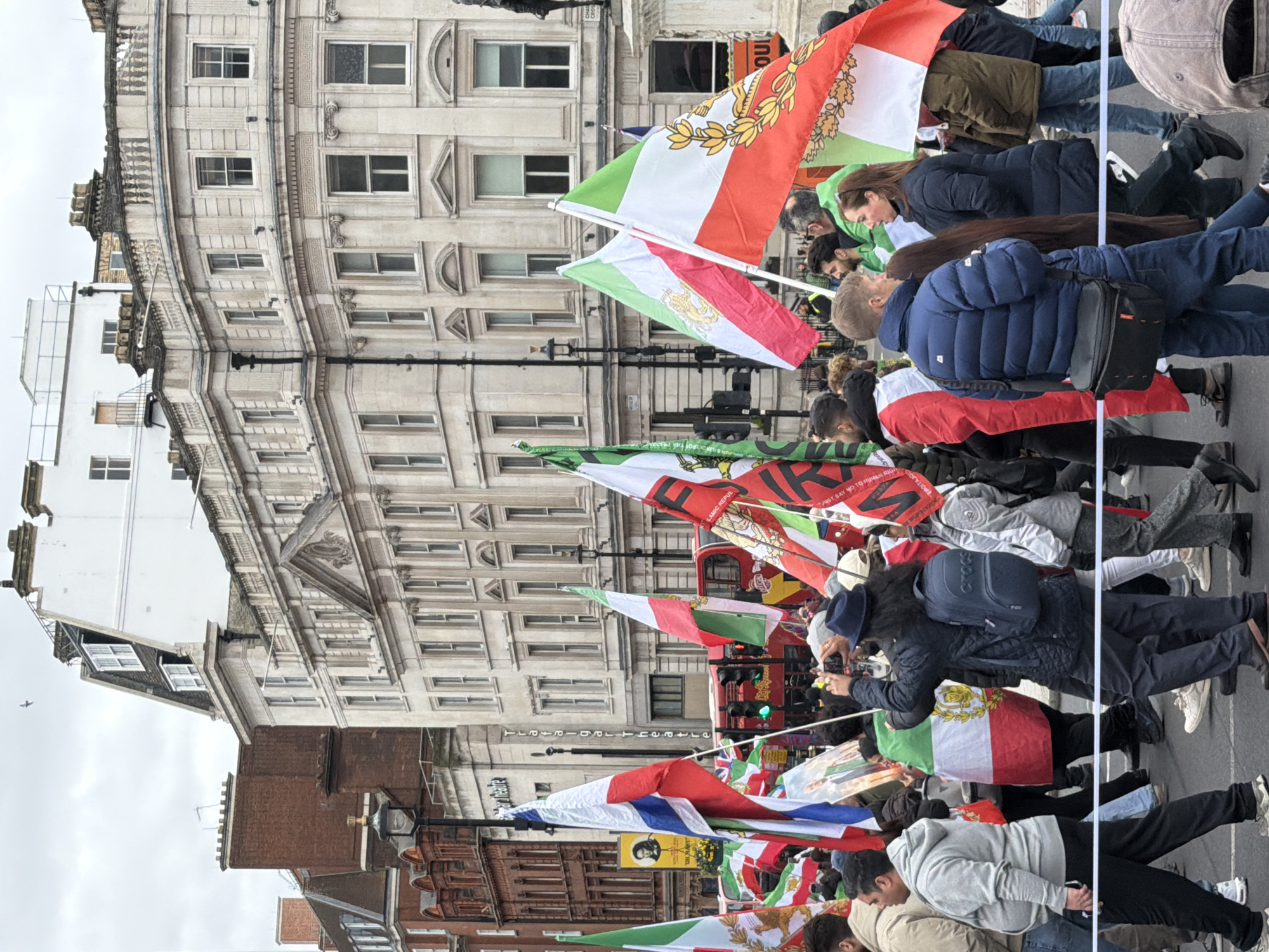 A crowd of people is seen in what appears to be a protest or demonstration in London, United Kingdom. Many individuals are holding and waving flags with green, white, and red stripes, some bearing an emblem. The architecture in the background suggests a city center location. The buildings feature multiple windows, ornate details, and a mix of light-colored stone and brick facades. A red double-decker bus is partly visible. The sky is overcast.