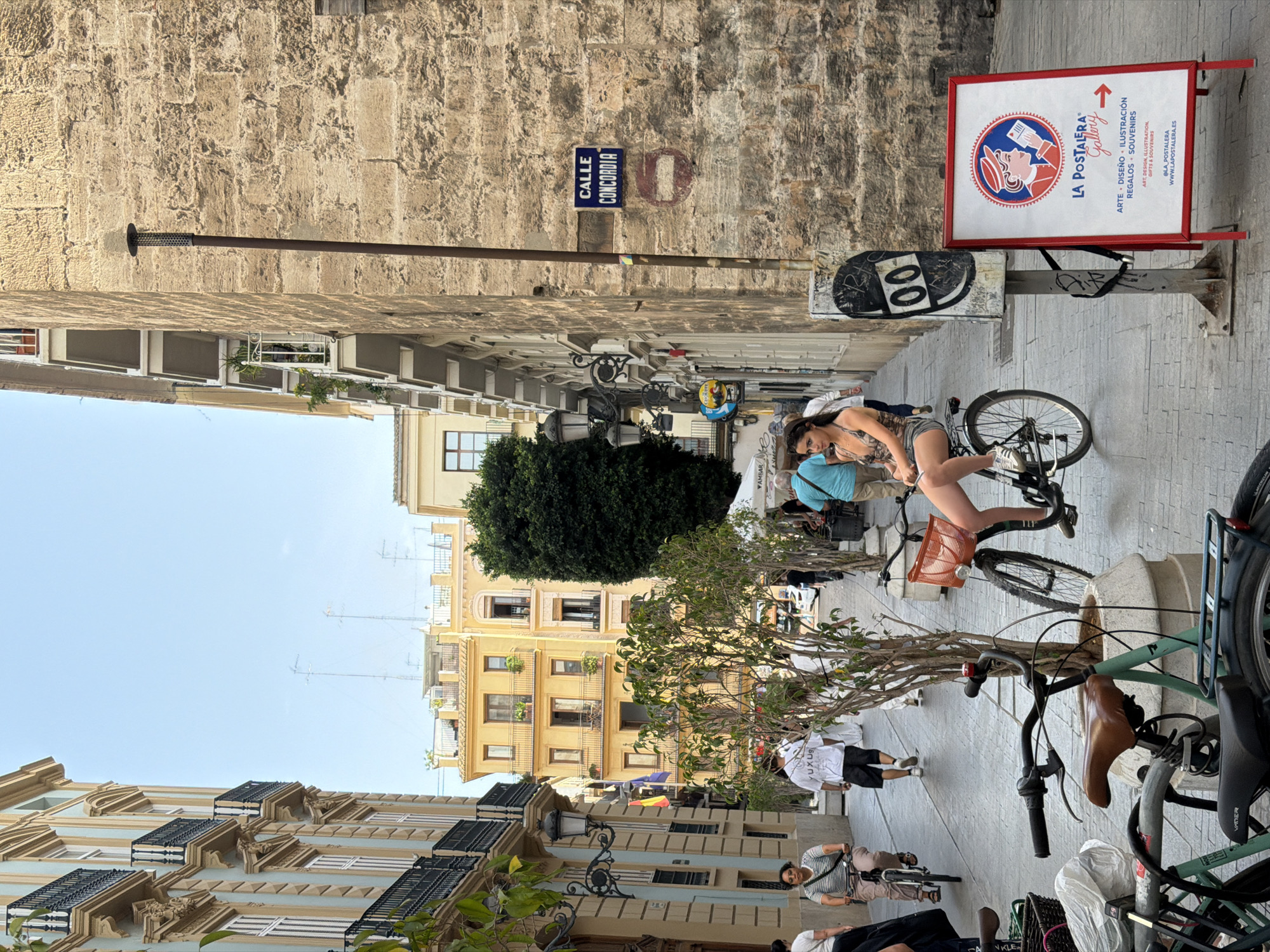 The image depicts a narrow cobblestone street in a European city, likely Spain given the signage. The street is lined with multi-story buildings featuring balconies and ornate architecture. On the right side of the street, a weathered stone building dominates the foreground, with a street sign reading "CALLE CONCORDIA." A prominent red and white A-frame sign for "LA POSTALERA Gallery" is positioned at the base of the building.
Several people are visible navigating the street. In the center foreground, a young woman is seated on a bicycle, looking towards the left of the frame. She is wearing a patterned top and denim shorts. In the background, various individuals are walking and cycling. One person on a bicycle is seen in the mid-ground to the left, wearing a white t-shirt and black shorts. Further back, other pedestrians are moving along the street, some partially obscured by planters with trees.
The environment appears to be a pedestrian area, possibly a market or shopping district, with trees in planters interspersed along the sidewalk. The sky above is a clear blue. The overall scene captures a lively moment in an urban setting during daylight hours.