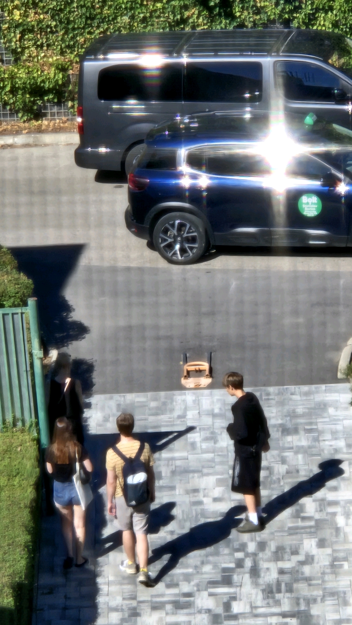 The image shows a scene from a high angle, looking down on a paved area adjacent to a hedge and a green metal fence. Two dark-colored vehicles, a van and a smaller car, are parked on the paved surface. The smaller car is dark blue and has a green circular sticker with white text on its side. In the foreground, four individuals are walking on the paved area. Two are a woman and a man walking away from the camera. The woman is wearing a black top and denim shorts, carrying a white tote bag. The man is wearing a striped t-shirt, shorts, and a backpack. Two other individuals are visible, one partially obscured by the man in the striped shirt and the other walking away from the camera. One of the men is wearing a black t-shirt and shorts, and appears to be standing. The environment suggests a sunny day with strong shadows cast by the people and objects. The paved surface is made of rectangular stones arranged in a repeating pattern. The hedge appears to be dense and green, bordering the paved area. The image quality is affected by glare from the sun, particularly on the vehicles. The location is identified as Prešov, Slovakia.