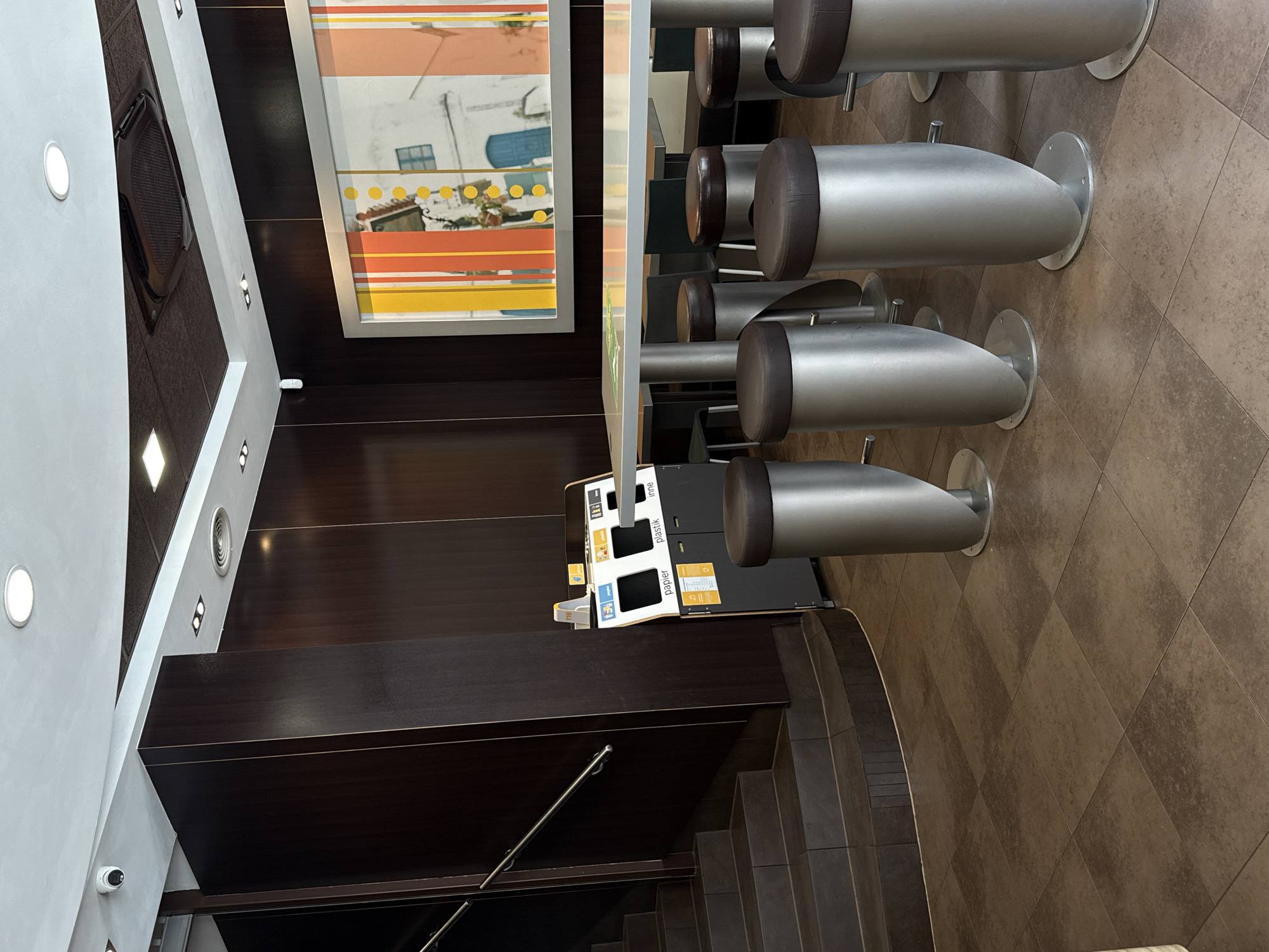 The image shows an interior view of a McDonald's restaurant in Łódź, Poland. The scene features a row of silver bar stools with dark brown seats, a recycling station with compartments for "papier" (paper) and "plastik" (plastic), and a staircase with dark brown wooden steps. The walls are dark brown, and the floor is tiled in a brown checkerboard pattern. A large, colorful abstract artwork hangs on the wall, depicting a white building with blue doors and windows, set against a background of orange and yellow stripes. Recessed lighting is visible on the ceiling. A security camera is mounted on a white wall on the left side of the image. The restaurant appears to be empty, with no people visible. The overall impression is one of a clean, modern fast-food establishment. The lighting suggests it is daytime.