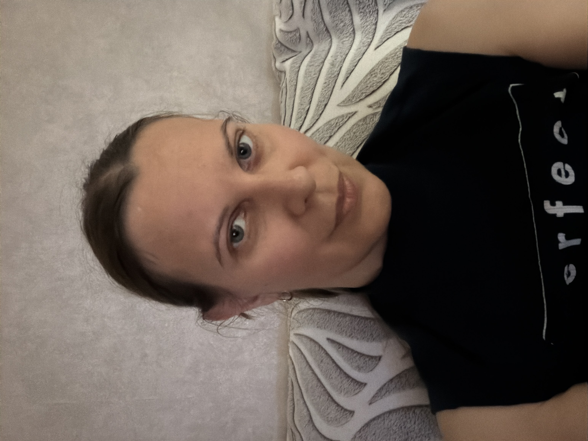 A frontal, medium-close-up photograph shows a woman with brown hair pulled back, fair skin, and light blue eyes. She is wearing a dark sleeveless shirt with white text and graphics. She is leaning back on a patterned cushion. The background is a textured, light-colored wall. The image was taken indoors.