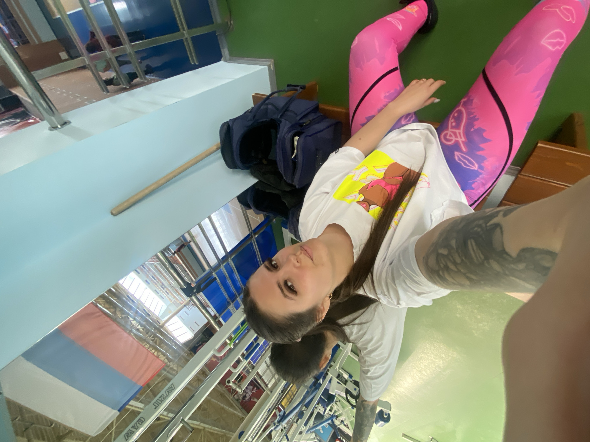 The image shows a woman taking a selfie inside a building in the city of Chaikovsky, Russia. The woman is wearing pink leggings with a black stripe and a white t-shirt with a cartoon image. She has dark hair and appears to have tattoos on her arms. In the background, there is a bag, a railing, and a Russian flag. The building appears to be some type of sports facility.