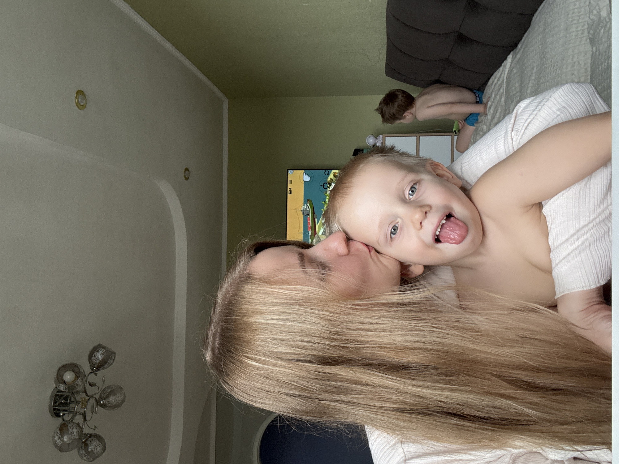 A photograph shows a woman and a child, possibly in a residence. The woman, with long blonde hair, is kissing the child on the cheek. The child, who has light hair and blue eyes, is sticking out his tongue. The room also contains a television that is on and playing a game, furniture, and what appears to be another child in the background. The location is Chaikovsky, Russia.