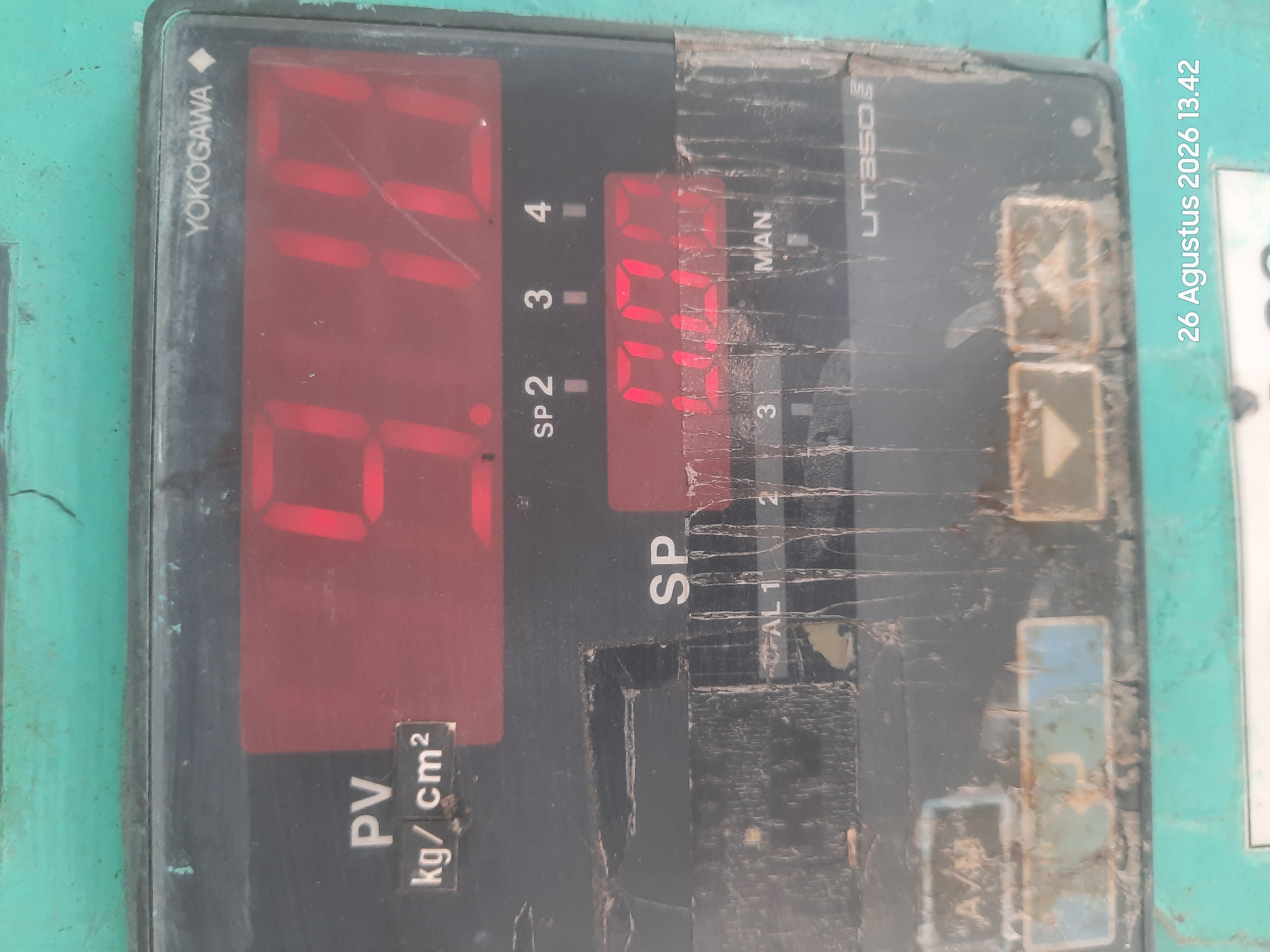 The image shows a close-up of a digital display panel, likely from industrial equipment, with the brand name "YOKOGAWA" visible at the top. The display shows red digital numbers and text. The top display shows "9.10" with "PV" and "kg/cm²" labels to its left. Below this, another display shows "0.00". Several labels such as "SP", "MAN", "AL 1", "2", and "3" are also visible. The panel is mounted on a teal-colored surface. A timestamp "26 Agustus 2026 13.42" is present at the bottom right corner of the image. The panel appears worn and dirty, with scratches and debris on its surface. The context of Gending, Indonesia is not directly evident from the image itself.