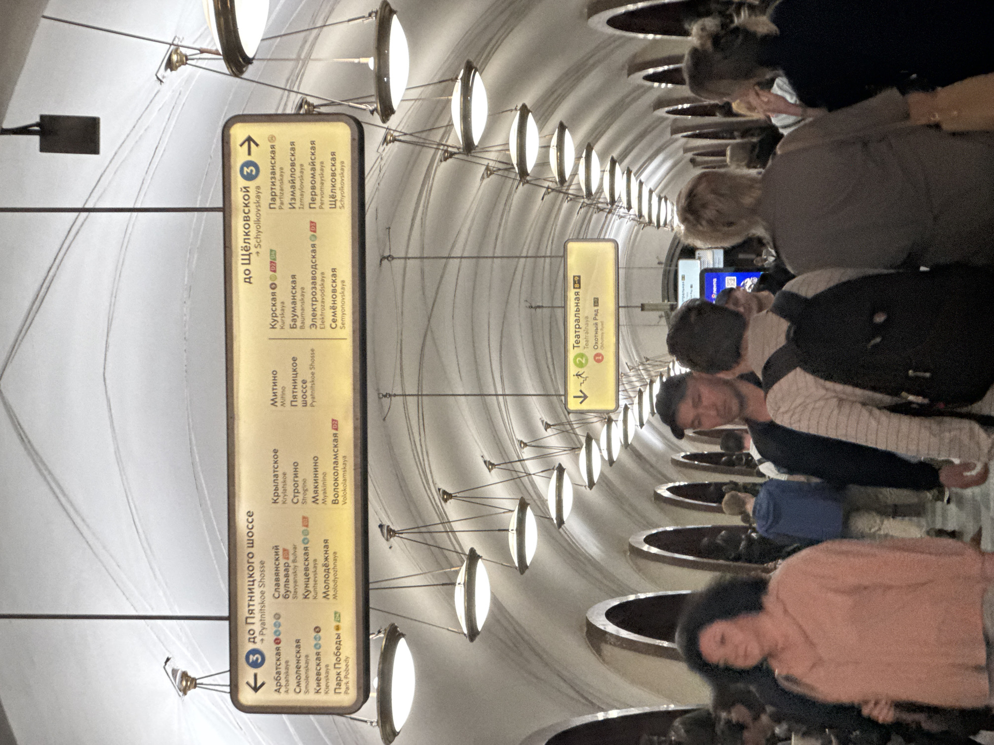 The image shows a Moscow Metro station. A large yellow sign with Cyrillic text, indicating metro lines and destinations, hangs from the ceiling. The sign lists stations such as "Арбатская," "Славянский бульвар," "Кунцевская," "Молодёжная," "Парк Победы," "Крылатское," "Строгино," "Мякинино," "Волоколамская," "Пятницкое шоссе," "Щёлковская," "Партизанская," "Измайловская," "Первомайская," "Курская," "Бауманская," "Электрозаводская," and "Семёновская." Another smaller yellow sign with Cyrillic text and icons indicates "Театральная" and "Охотный ряд." The station features arched ceilings and rows of decorative hanging lights. Several people are visible, walking in different directions. Some are in the foreground, while others are further back in the station, blurred by motion. The overall atmosphere is that of a busy, functional public transit space.