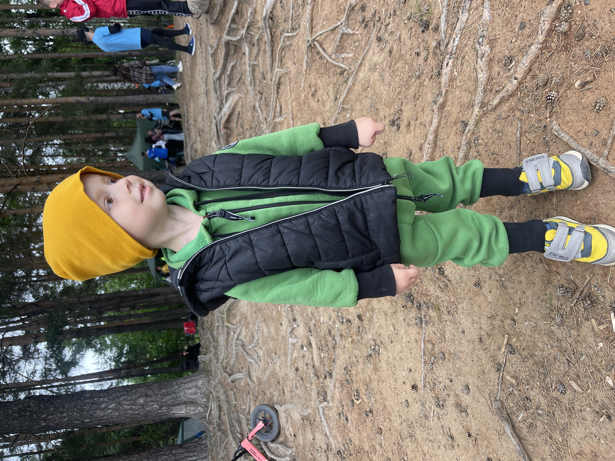 A young boy wearing a yellow beanie, a green hooded sweatshirt, a black puffer vest, and green sweatpants stands in a wooded area. He is also wearing black socks and grey and yellow sneakers. Behind him, other people are visible in the background, some standing near tents, suggesting an outdoor gathering or event. The ground is covered with sand, pine needles, pinecones, and exposed tree roots. The environment appears to be a forest or park area within or near the city of Chaikovsky, Russia, possibly during an outdoor festival or camping trip. A pink children's bicycle is visible to the left of the boy.