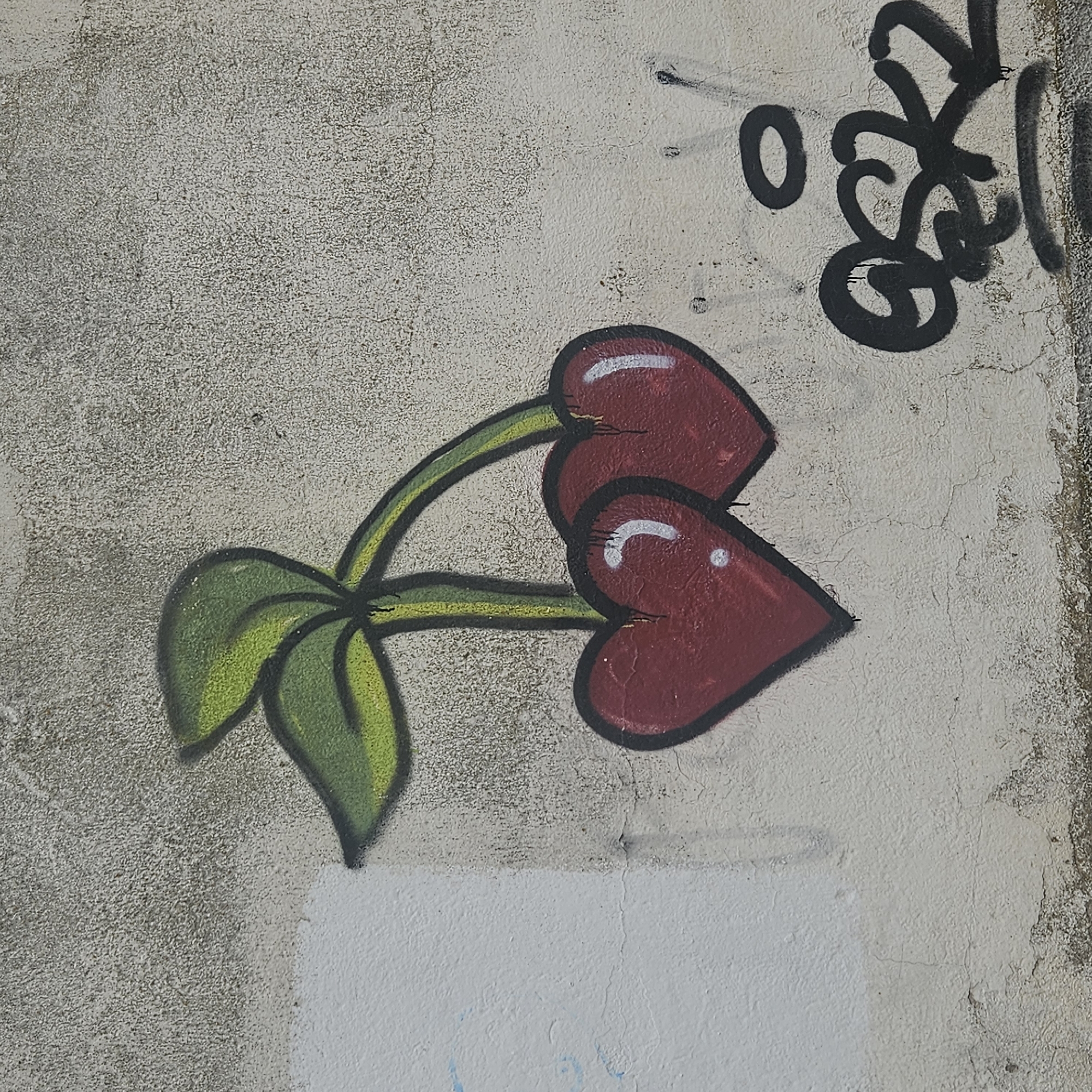 The image displays a section of a textured wall, likely concrete or stucco, located in Salt, Spain. A piece of graffiti, depicting two heart-shaped cherries, is centrally positioned. The hearts are rendered in deep red with thick black outlines, each featuring a prominent white highlight. They are connected by two green stems, also outlined in black and accented with lighter green, displaying paint drips. Two green leaves, outlined in black with lighter green accents, emerge from the stems. The wall surface is a mottled light gray to off-white, characterized by a rough, granular texture and numerous fine cracks. A vertical seam or division is visible on the left, separating a slightly smoother, whiter section from the coarser main wall. Faint blue markings are present on the far left. In the lower right quadrant, below the cherry graffiti, distinct black graffiti tagging, including a stylized letterform (possibly "OSK") and a detached black circle, is evident, also exhibiting paint drips. The bottom edge of the wall shows increased discoloration and wear.